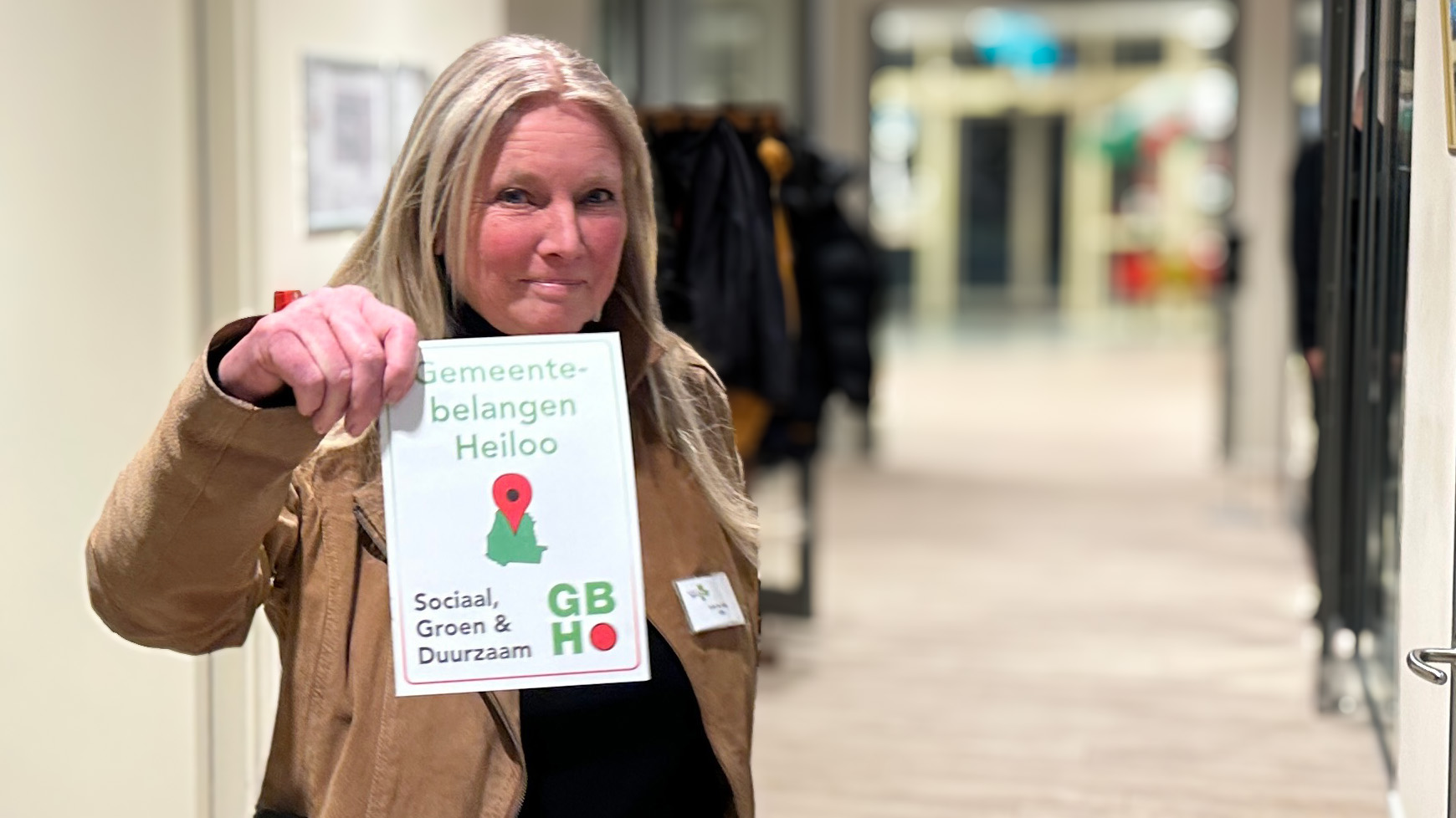 Vrouw houdt een bordje vast met de tekst "Gemeentebelangen Heiloo: Sociaal, Groen & Duurzaam", in een hal.