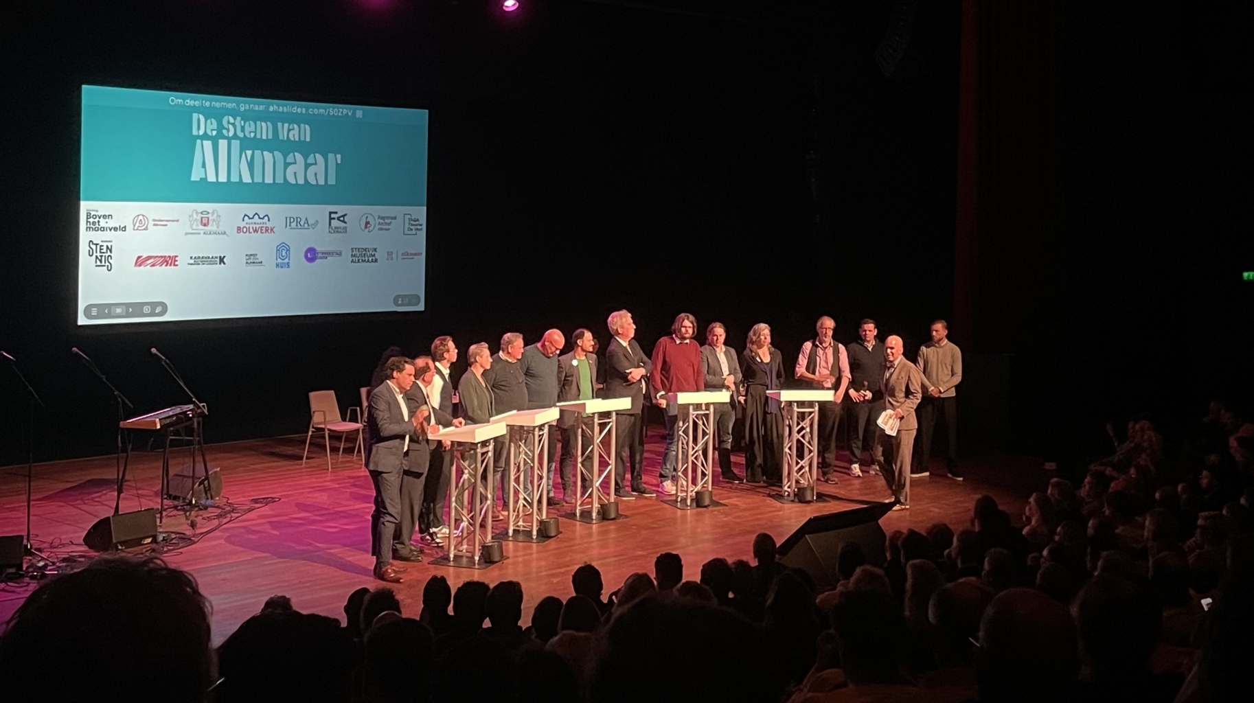 Mensen staan op een podium tijdens een evenement in een theater, met een scherm dat "De Stem van Alkmaar" toont.
