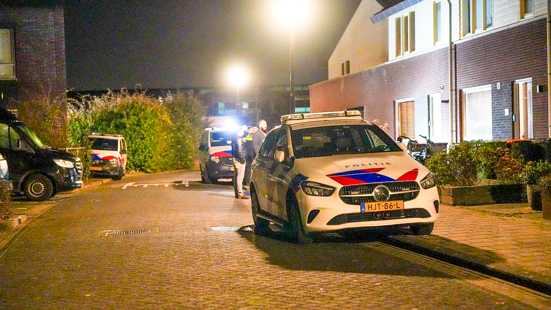 Politieauto's geparkeerd op een straat 's nachts, met meerdere mensen die naast een voertuig staan praten.