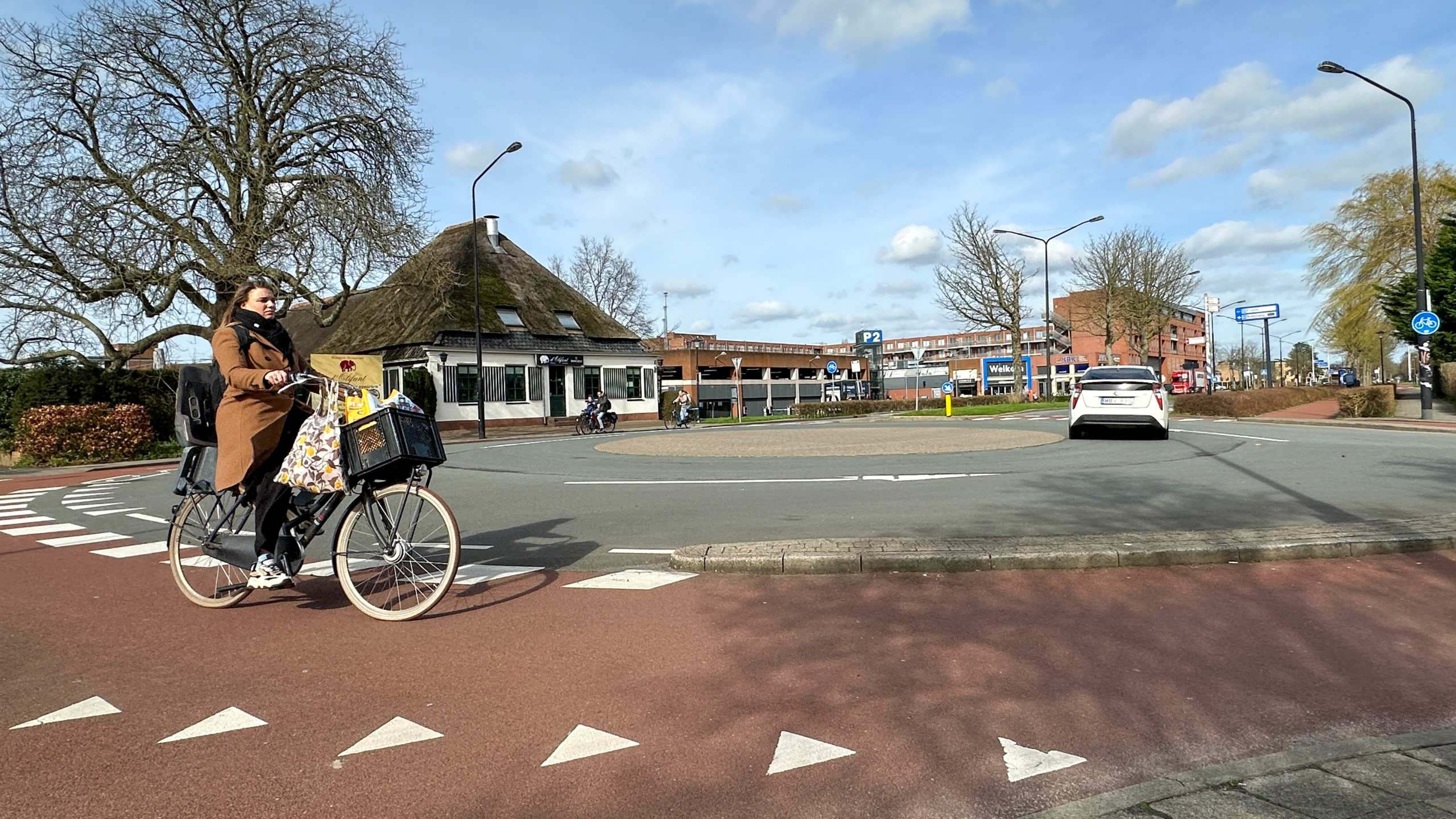 Vrouw fietst over een rotonde met een rieten dakgebouw en een auto op de achtergrond onder een blauwe lucht.