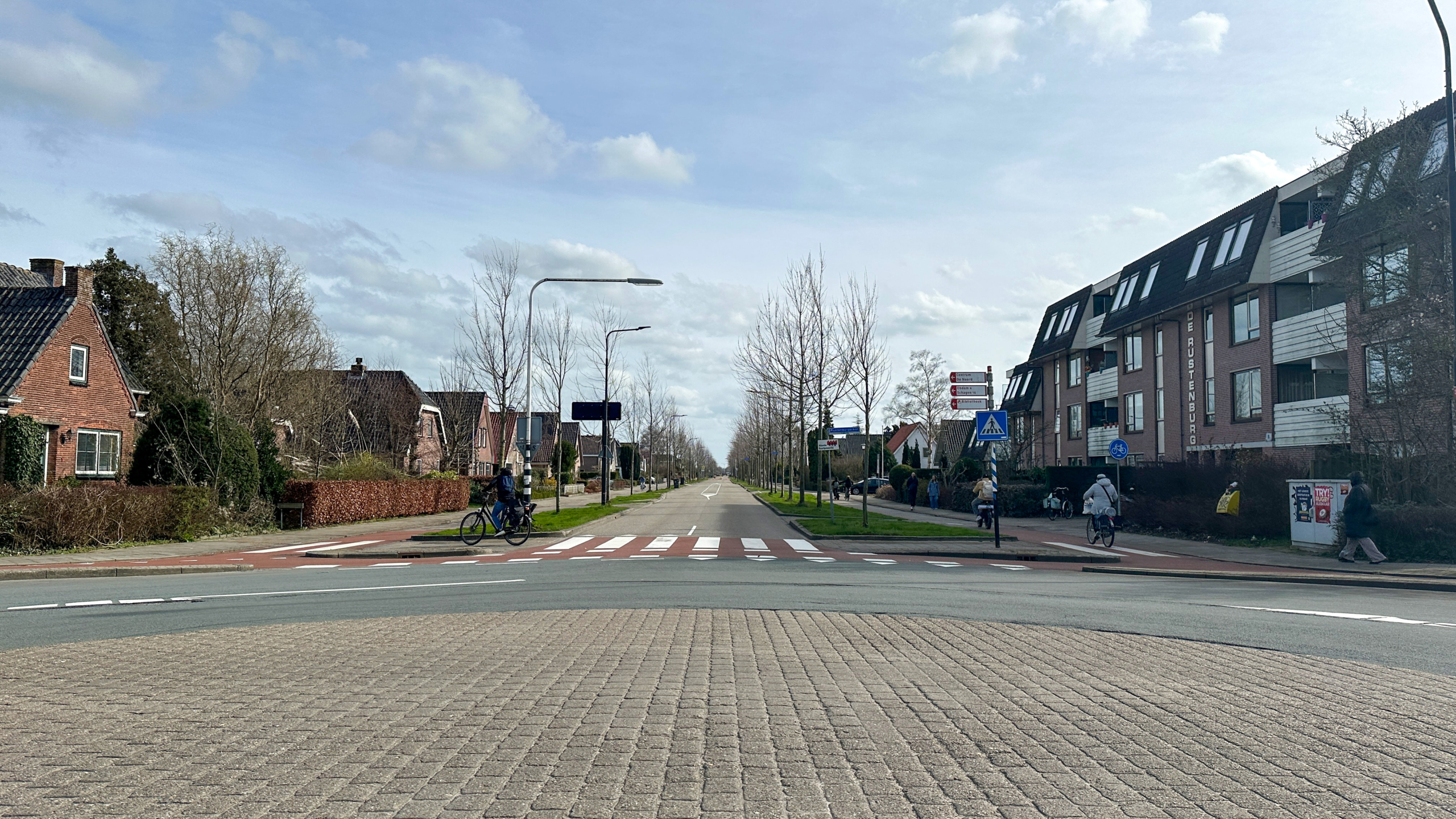 Straatbeeld met een zebrapad en fietspad, omgeven door huizen en appartementen, met enkele fietsers en voetgangers.