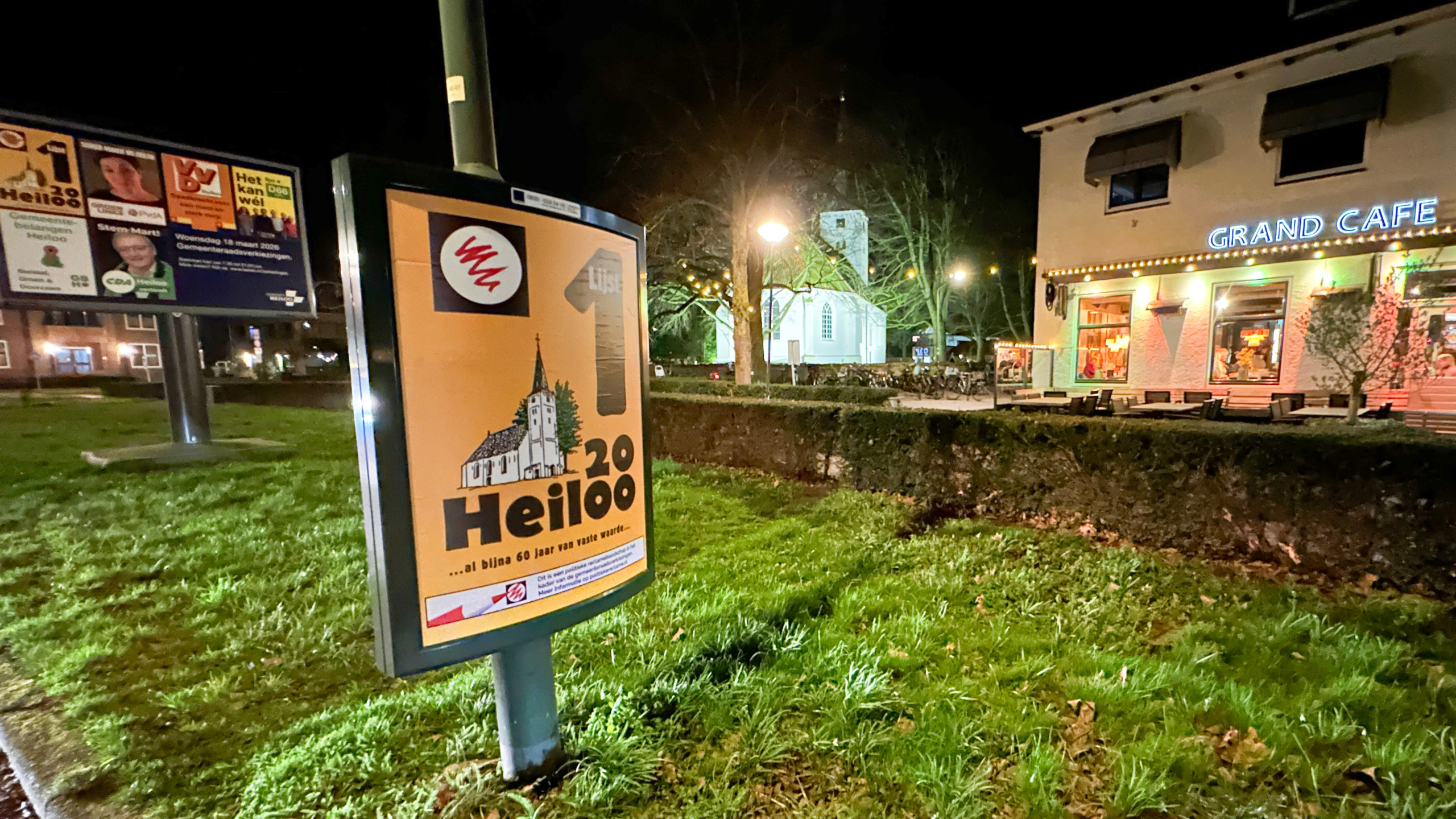 Verlichte verkiezingsposters in Heiloo met op de achtergrond een Grand Café en een verlichte kerk in de nacht.