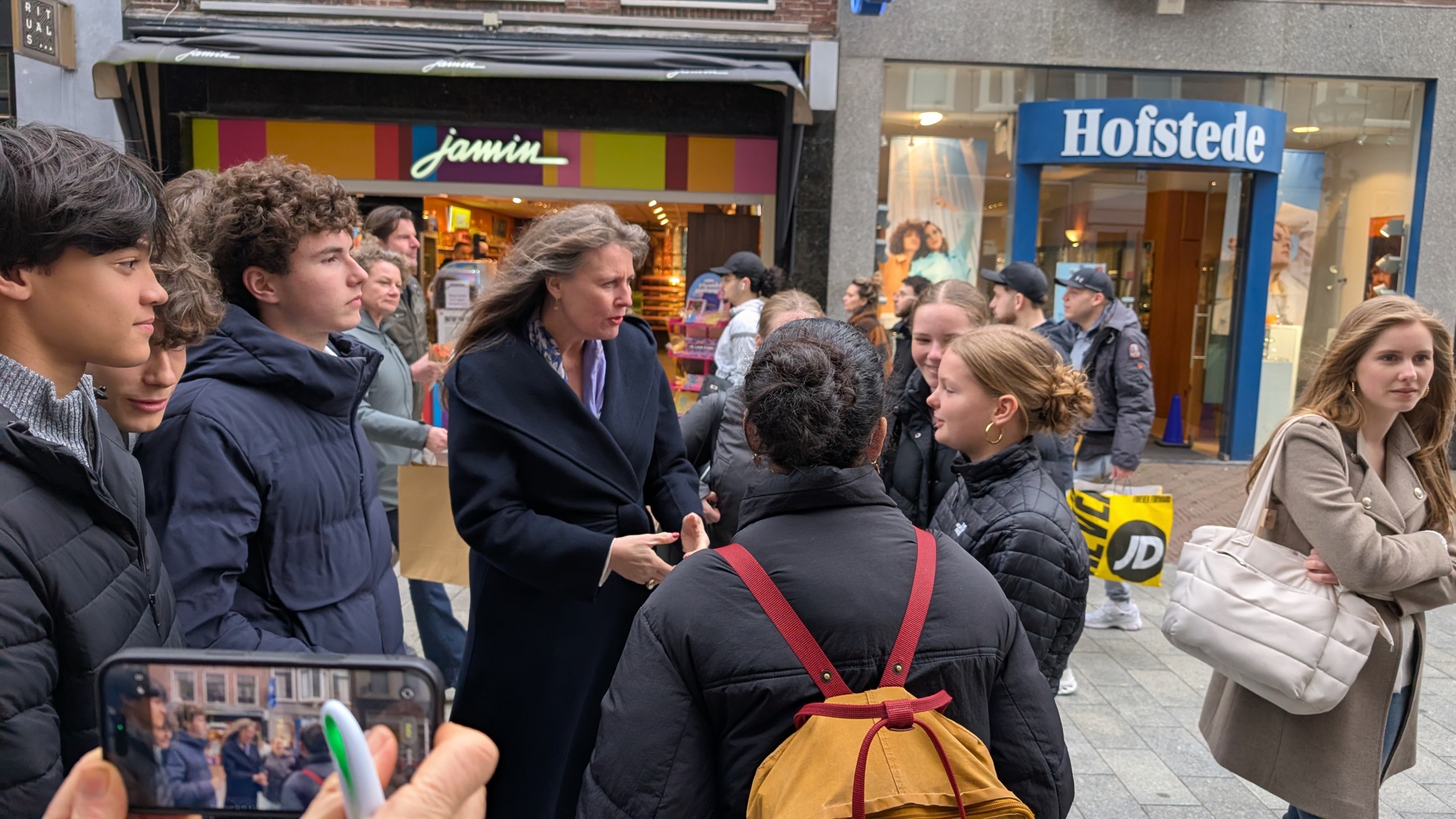 Mensen staan in een winkelstraat en praten met elkaar; op de achtergrond zijn winkels van Jamin en Hofstede te zien.