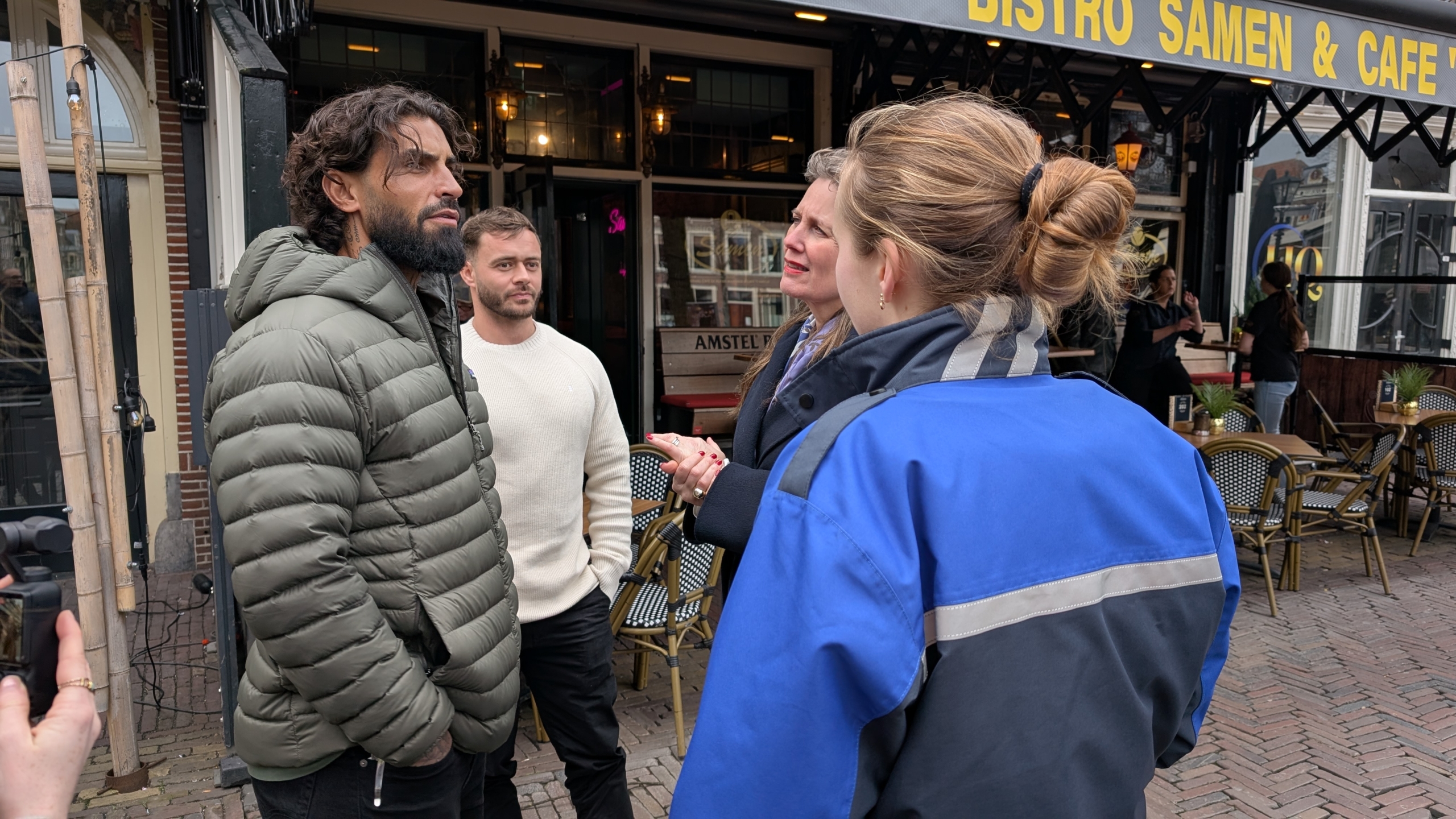 Mensen staan buiten bij een bistro terwijl een vrouw in een blauw jack met hen praat.