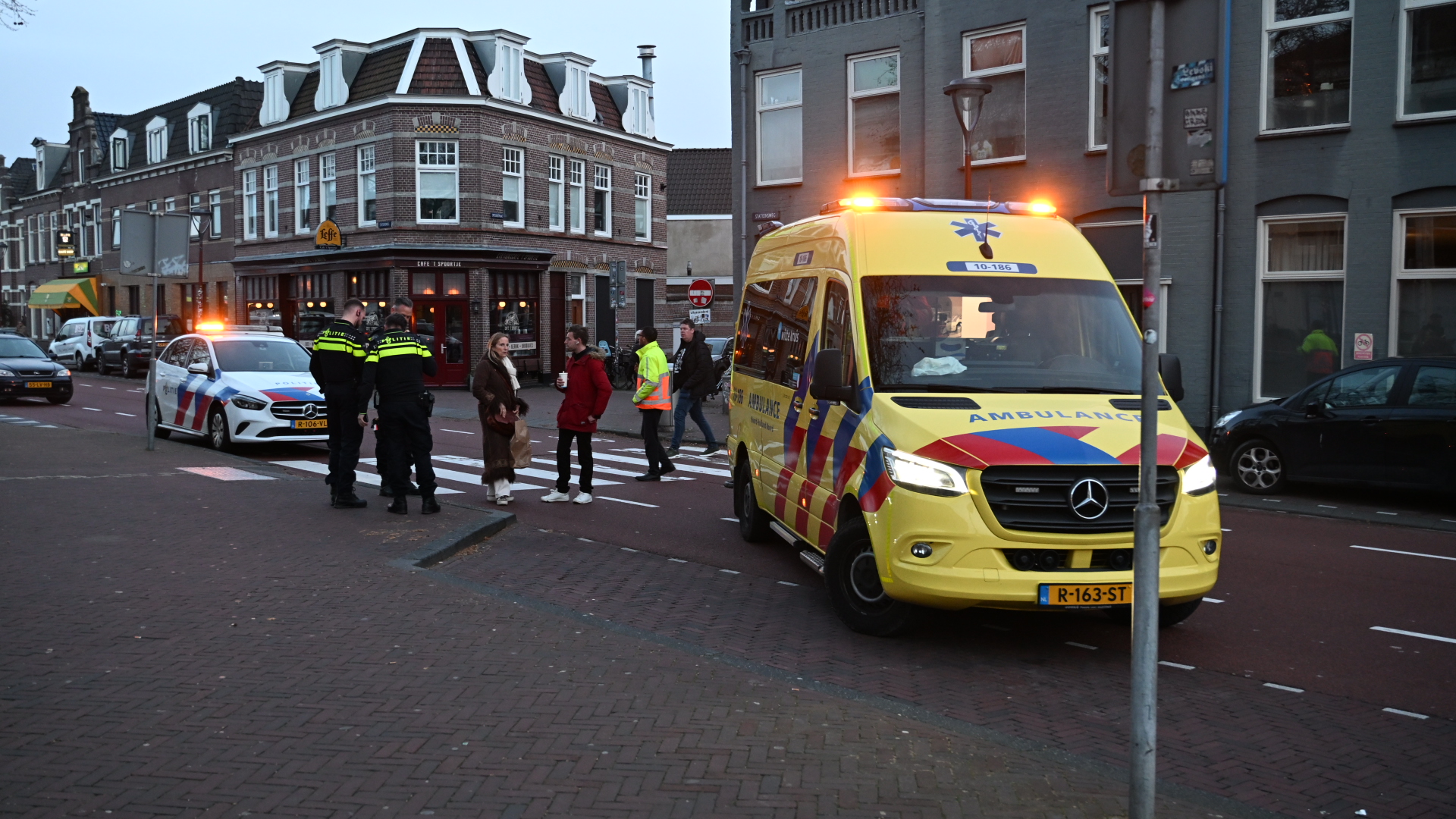 Een ambulance en politieauto staan met zwaailichten aan bij een kruising, omringd door omstanders en agenten.