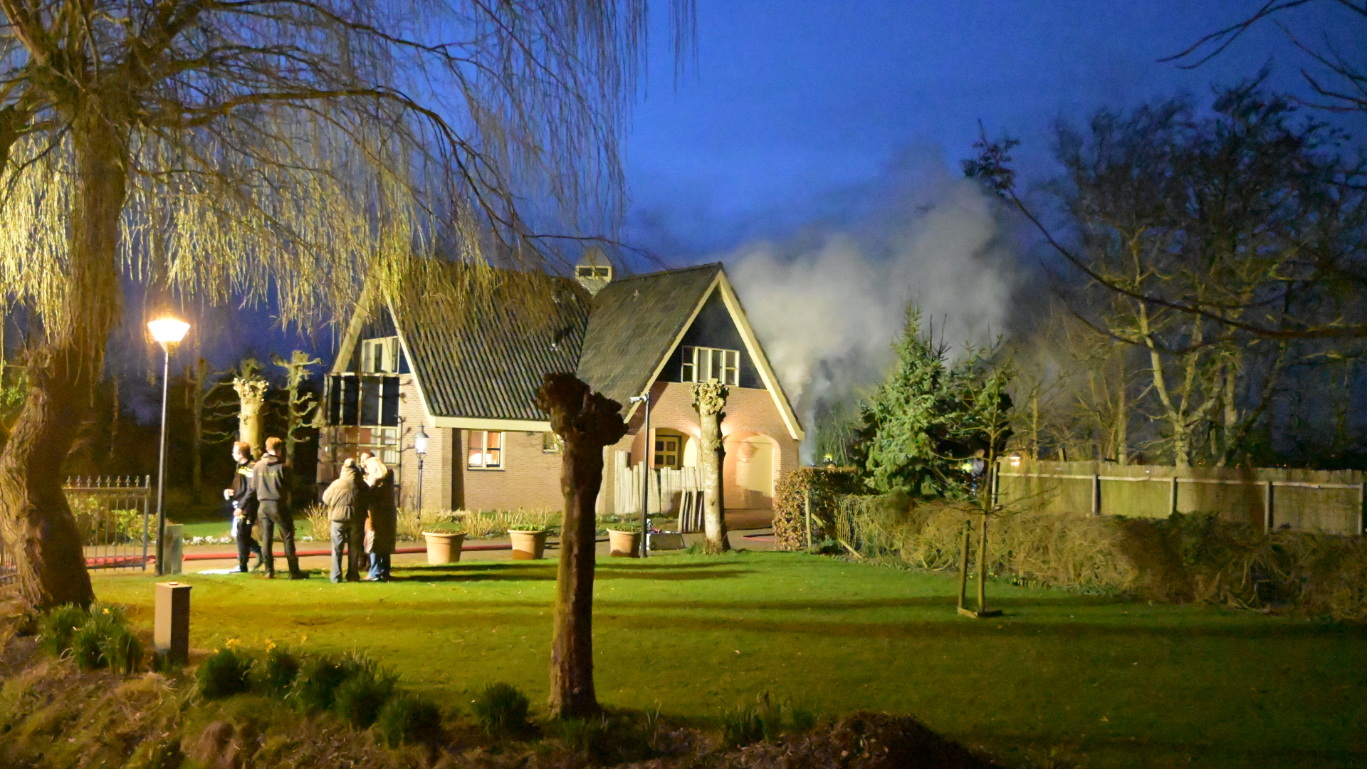 Een nachtelijk tafereel van een huis met rook die uit het dak komt, en een paar mensen die buiten staan.