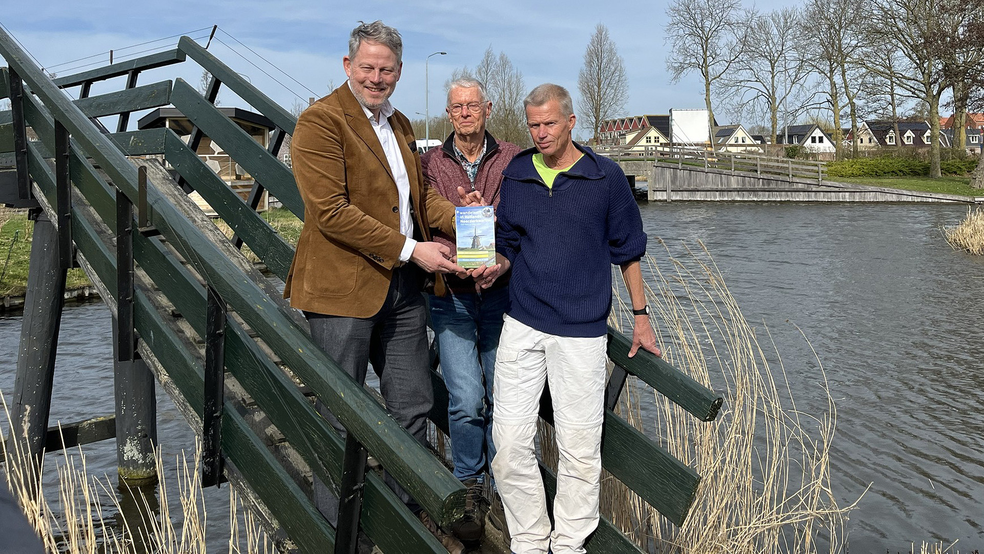 Drie mannen poseren op een houten brug naast een rivier terwijl ze een boek vasthouden met een afbeelding van een molen.