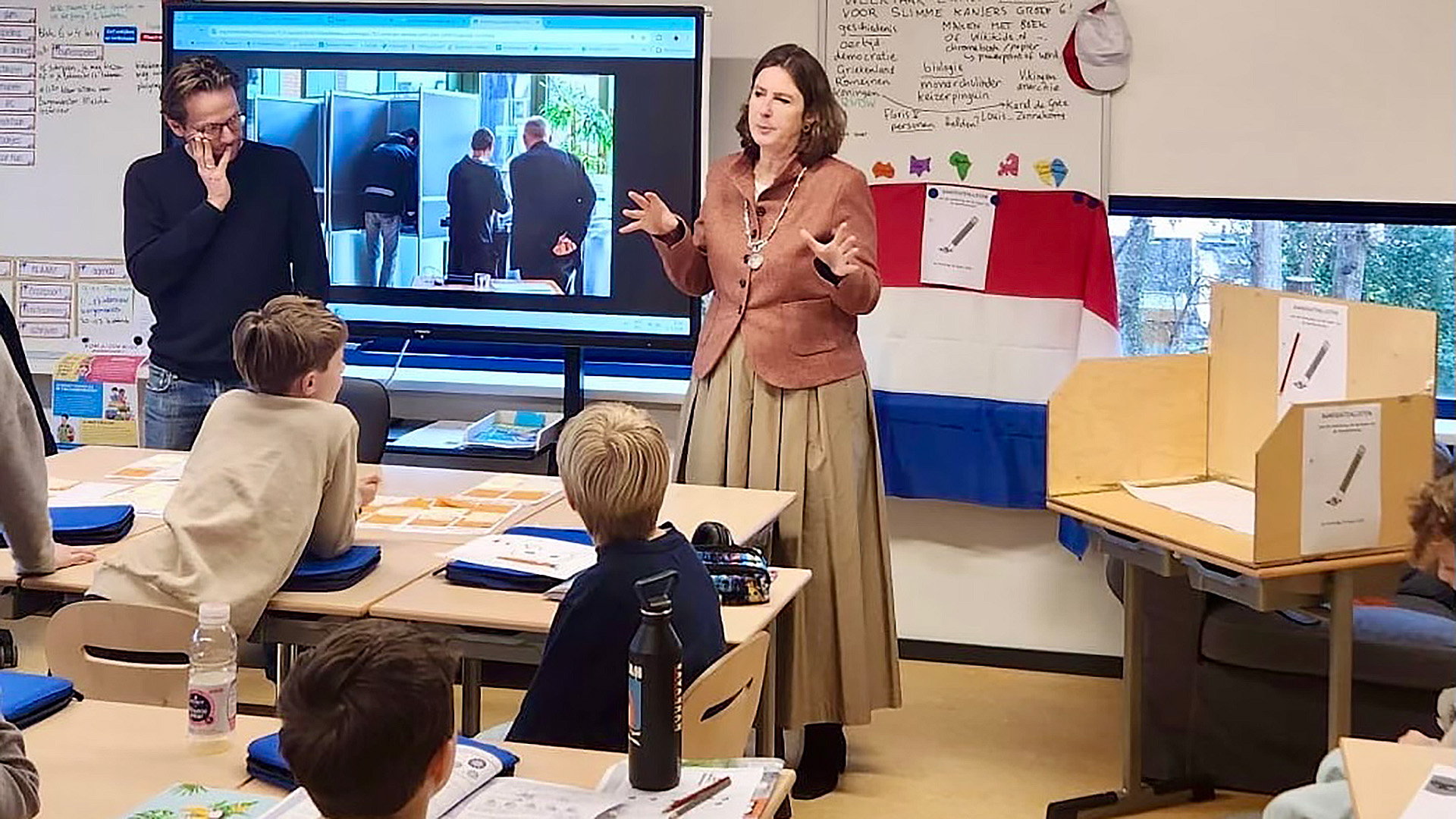 Een vrouw spreekt voor een klas met kinderen, er is een projectie op een scherm en een Nederlandse vlag hangt op de achtergrond.