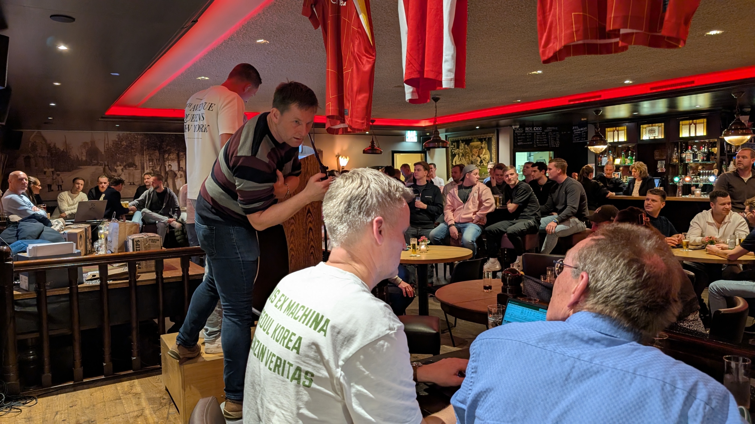 Een groep mensen in een druk café of bar setting, sommige zitten en anderen staan, met een man op een tafel die iets presenteert.