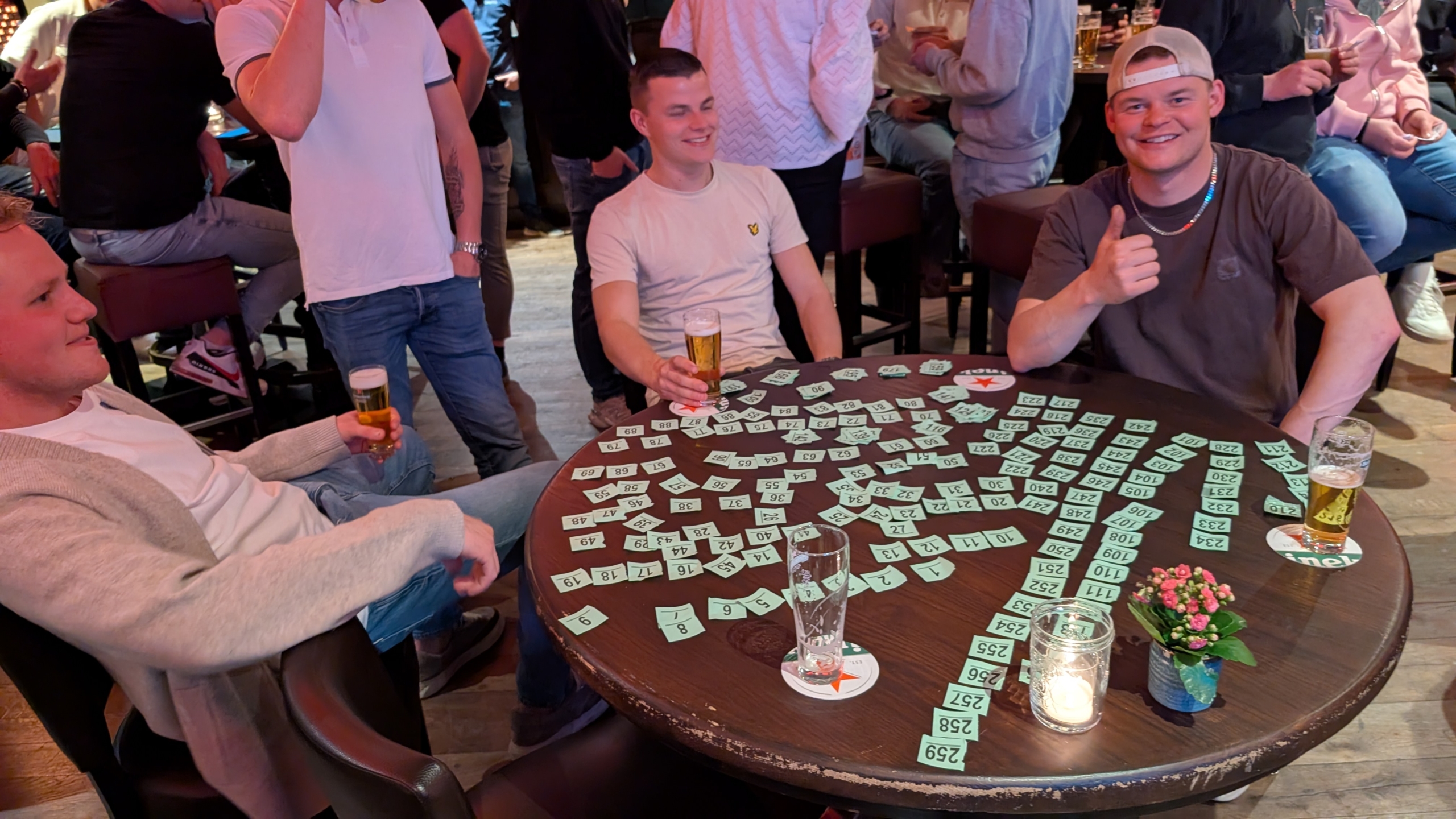 Drie mannen zitten aan een tafel vol met loten en bierglazen in een drukke bar; een van hen steekt zijn duim op.