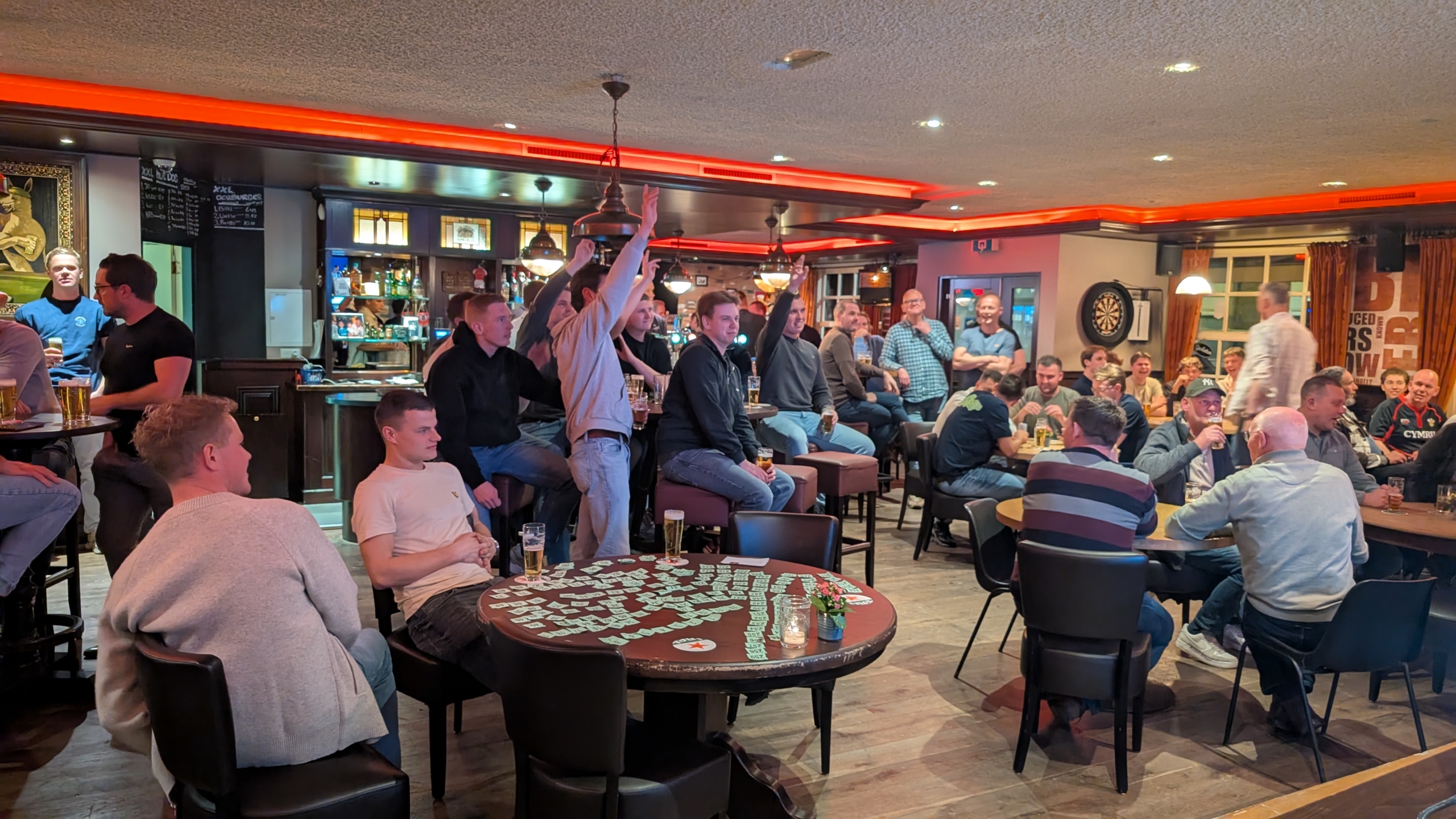 Mensen in een bar kijken naar iets buiten beeld, sommigen met handen omhoog, glazen bier op tafel.