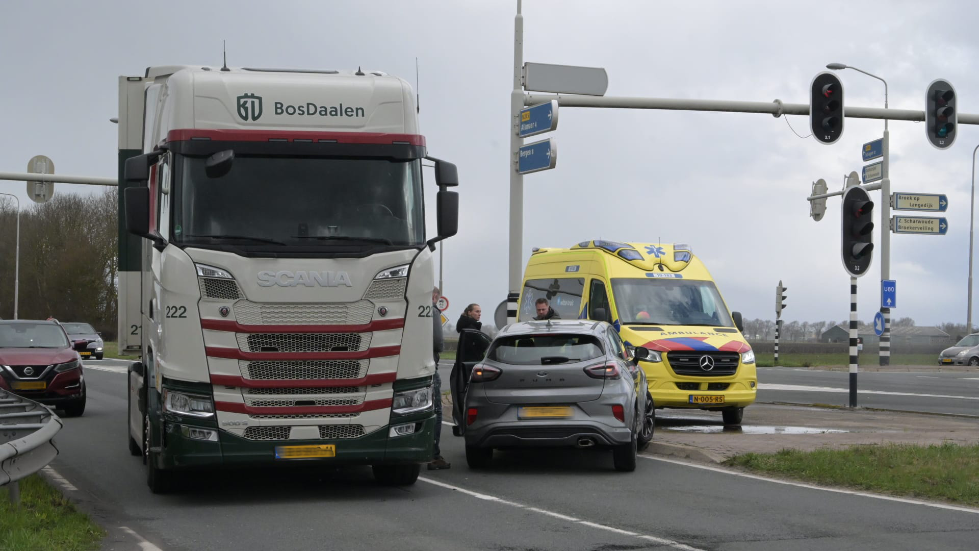 Een vrachtwagen, auto en ambulance staan stil bij een verkeerslicht. Mensen staan naast de voertuigen in gesprek.