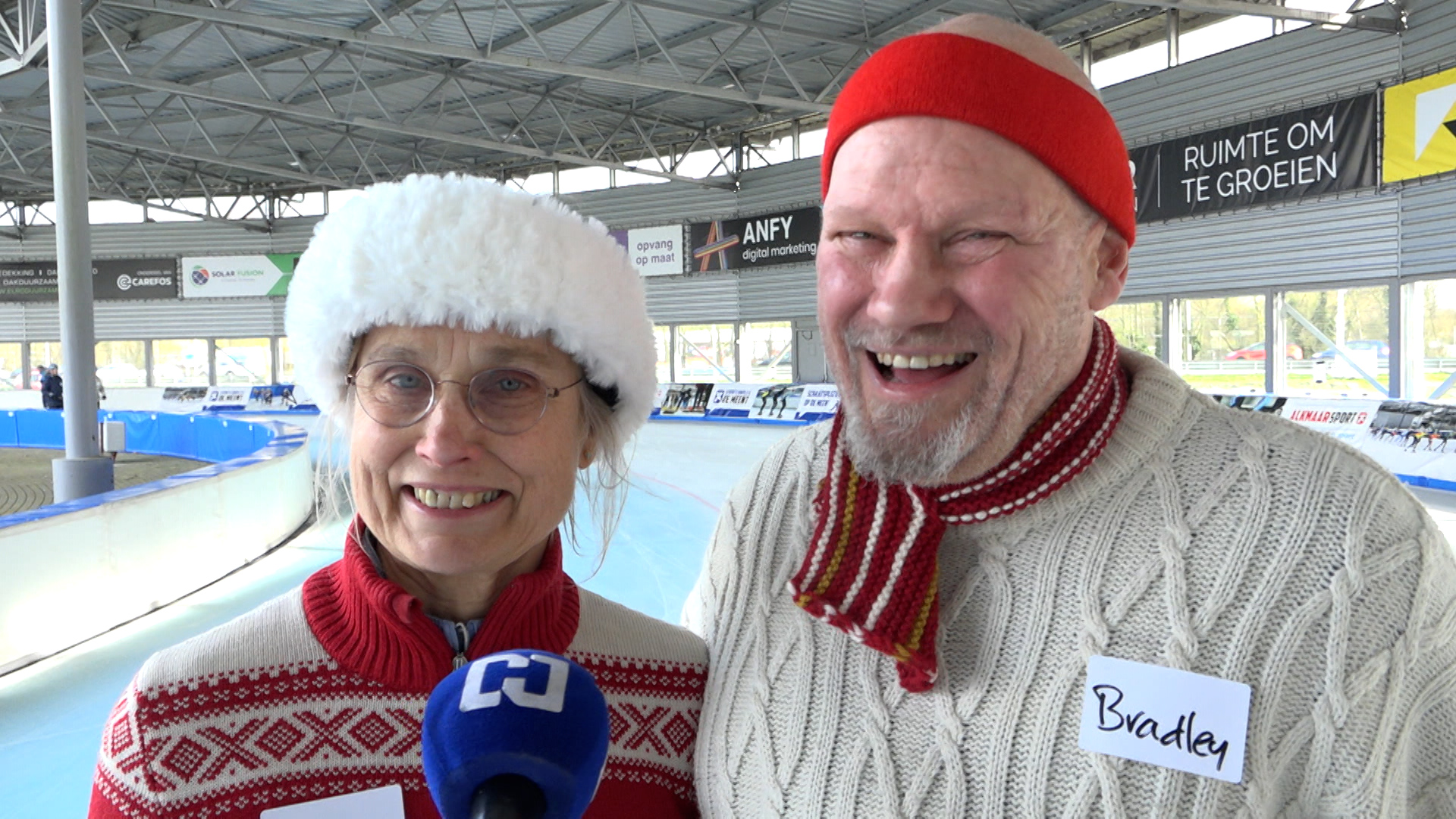 Twee lachende mensen in winterkleding bij een ijsbaan, met een microfoon voor hen.