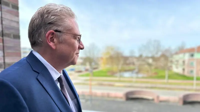 Man in een blauw pak kijkt uit over een rustige stedelijke omgeving met bomen en een waterpartij.