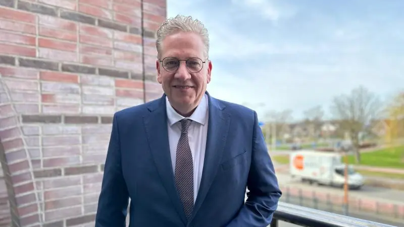 Man in een donkerblauw pak staat op een balkon met uitzicht over een weg en een grasveld.