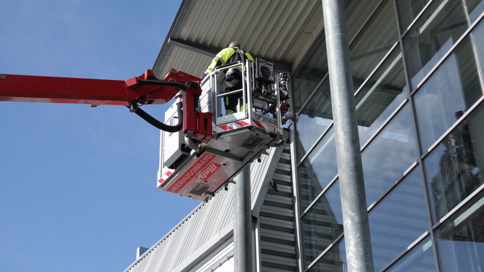 Persoon in een hoogwerker voert werkzaamheden uit aan een glazen gevel van een gebouw onder een blauwe lucht.