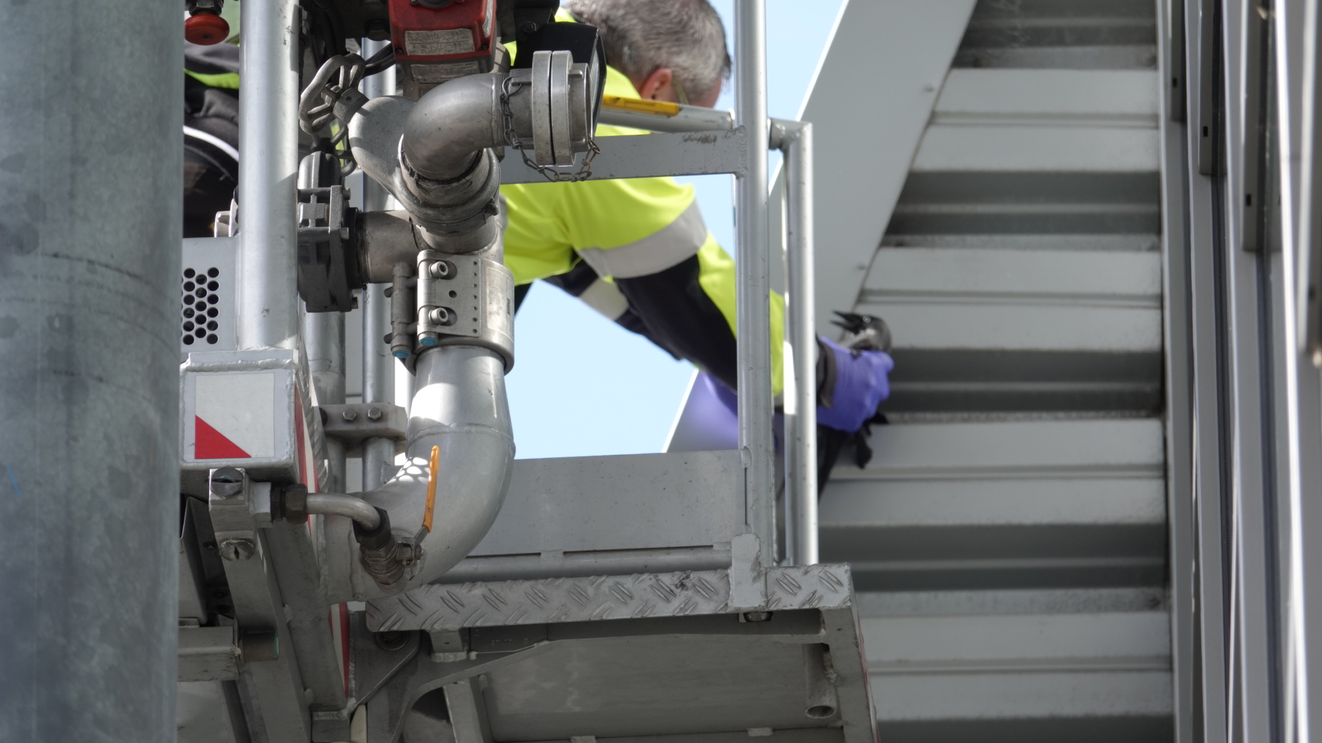 Een persoon in veiligheidskleding werkt op hoogte aan een metalen constructie met buizen en leidingen.
