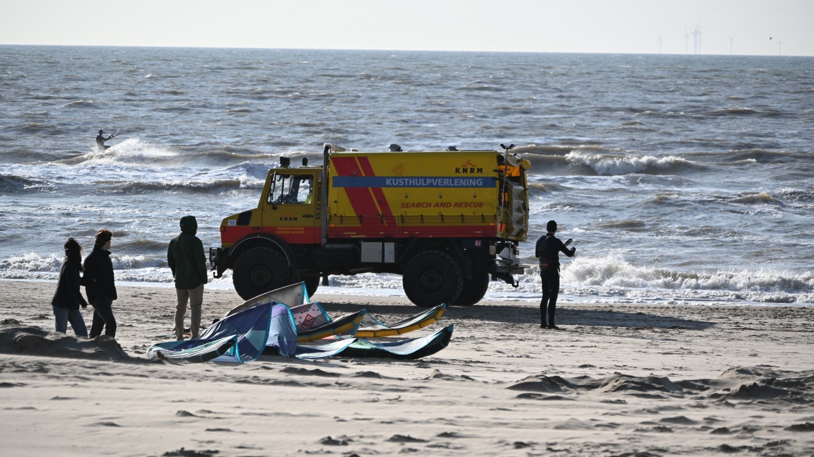 Kusthulpverleningsvoertuig van de KNRM op een strand met personen en een kitesurfer op zee op de achtergrond.
