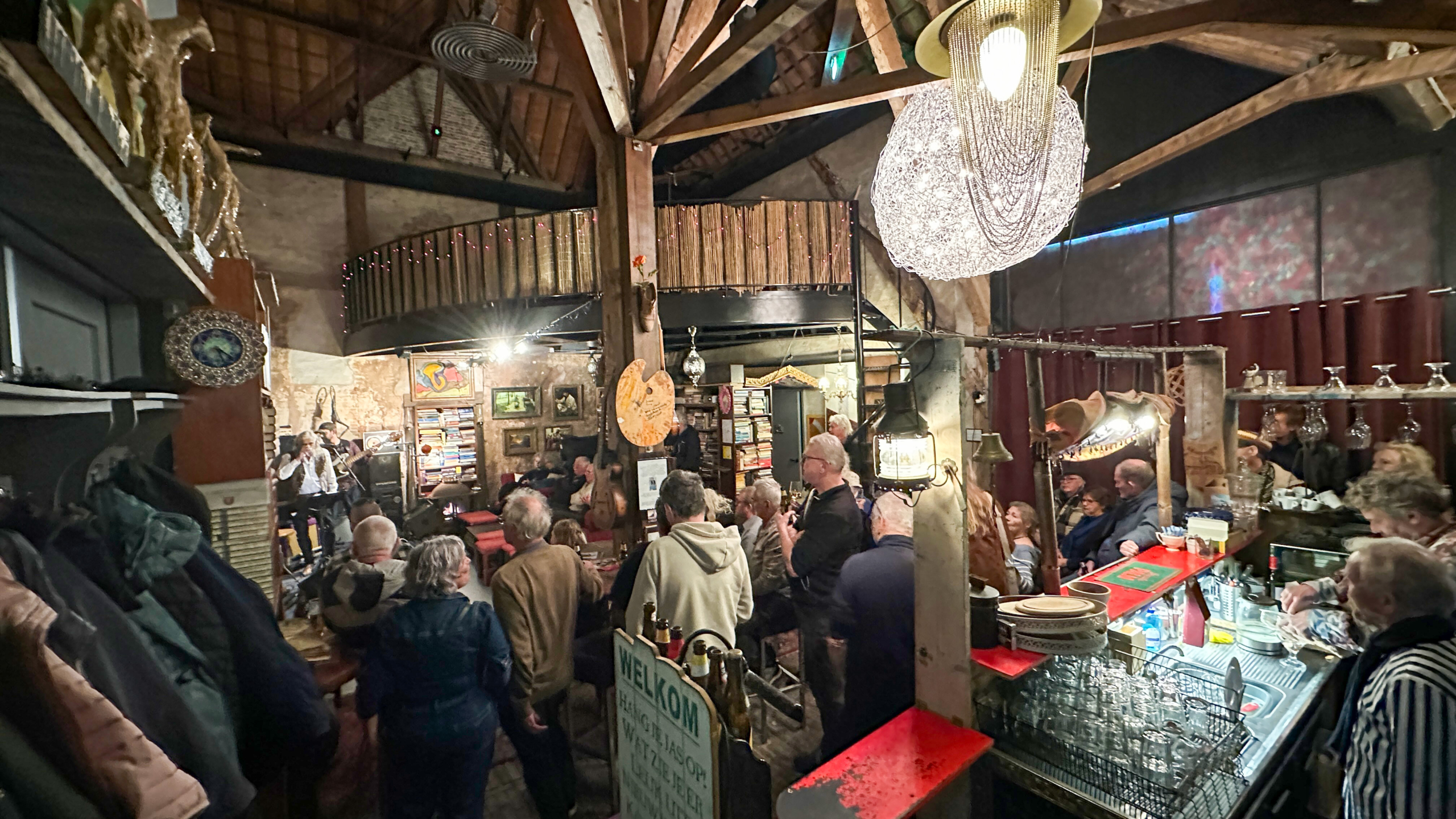 Mensen luisteren naar een muziekoptreden in een sfeervolle, rustieke bar met een houten interieur en gezellige verlichting.