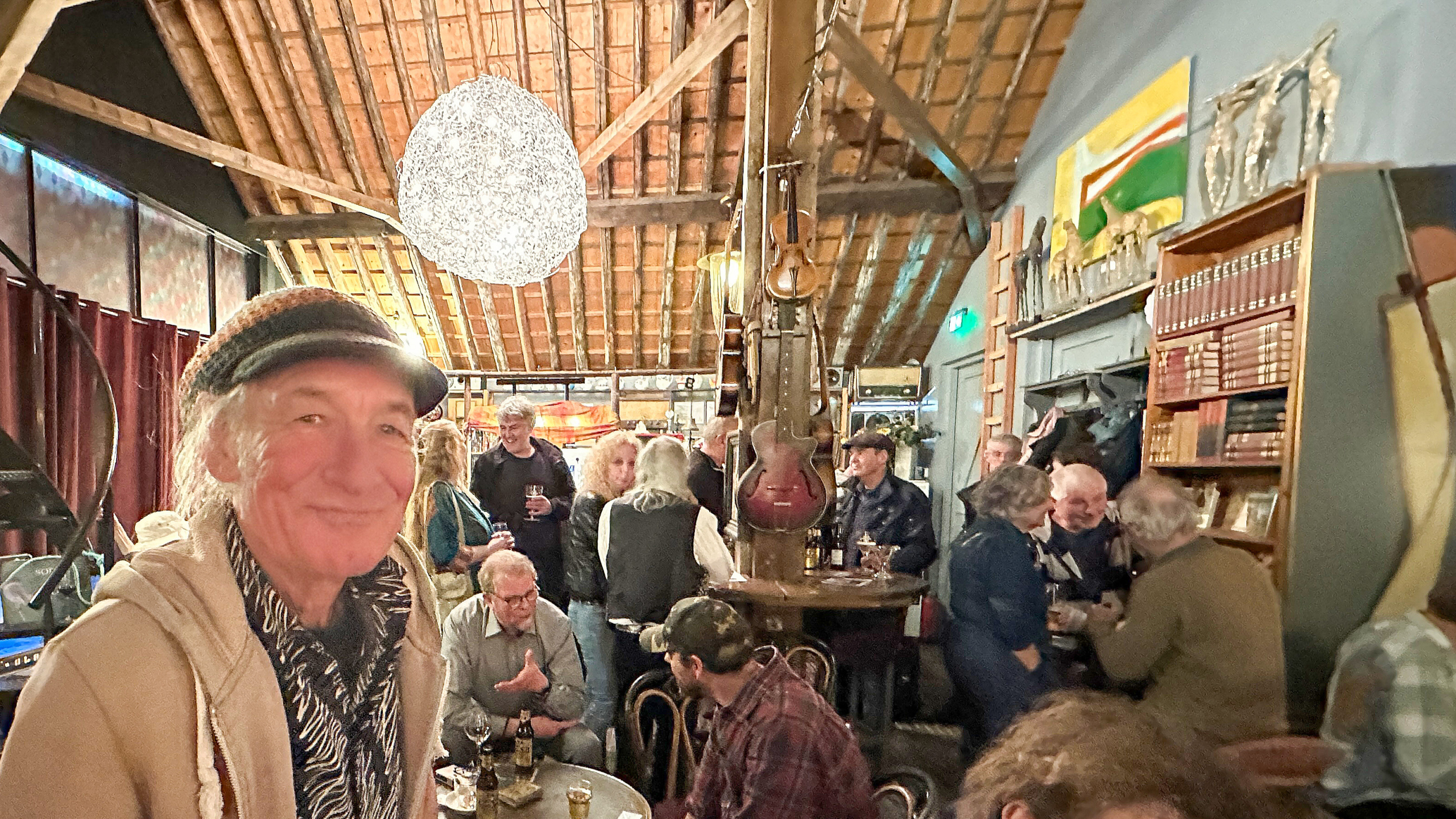 Een gezellige setting in een café met houten balkenplafond, een man op de voorgrond en groepen mensen die praten en drinken.