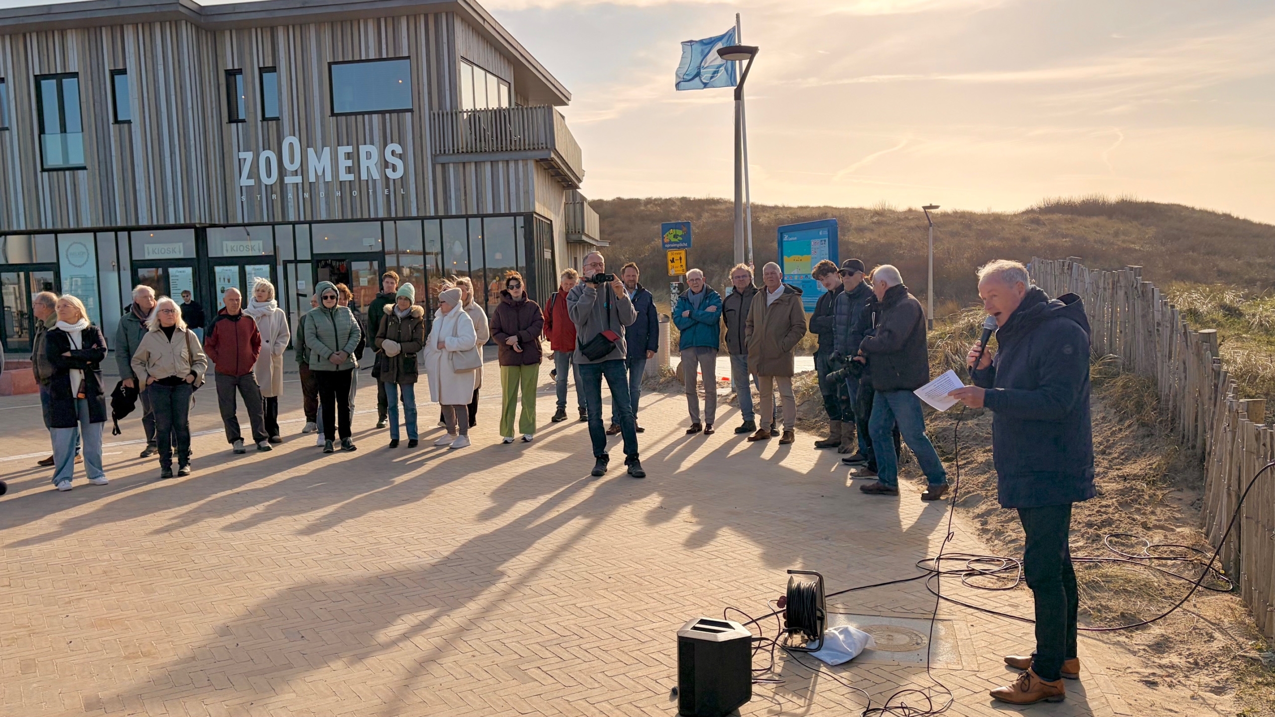 Een man spreekt voor een groep mensen buiten bij het Zoomers strandhotel, met een microfoon in de hand.