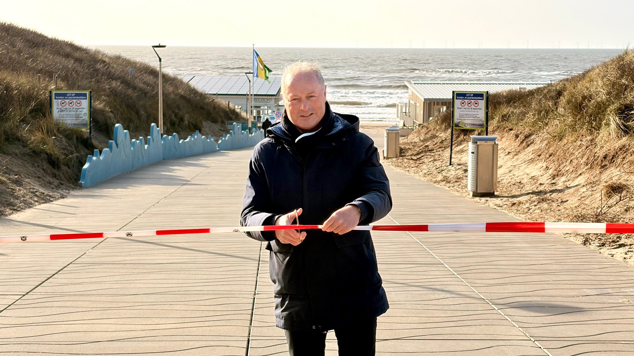 Een man knipt een lint door op een pad naar het strand, met de zee op de achtergrond.