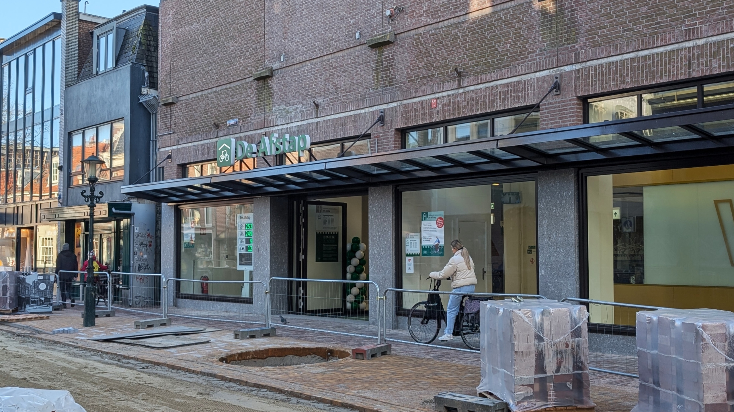Een vrouw met een fiets voor een modern gebouw met een bordje van "De Aftrap" en straatwerken op de voorgrond.