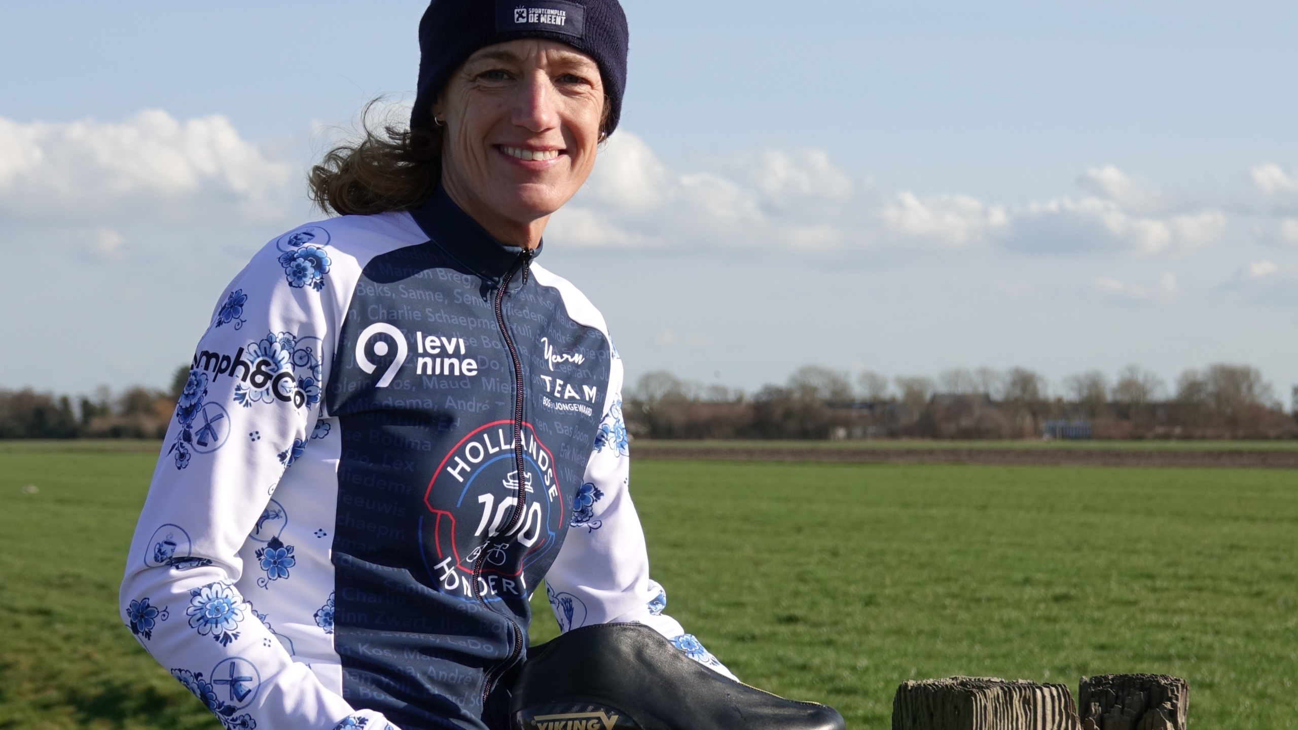 Vrouw in sportieve outfit met muts poseert met een schaats in een open veld onder een blauwe hemel.