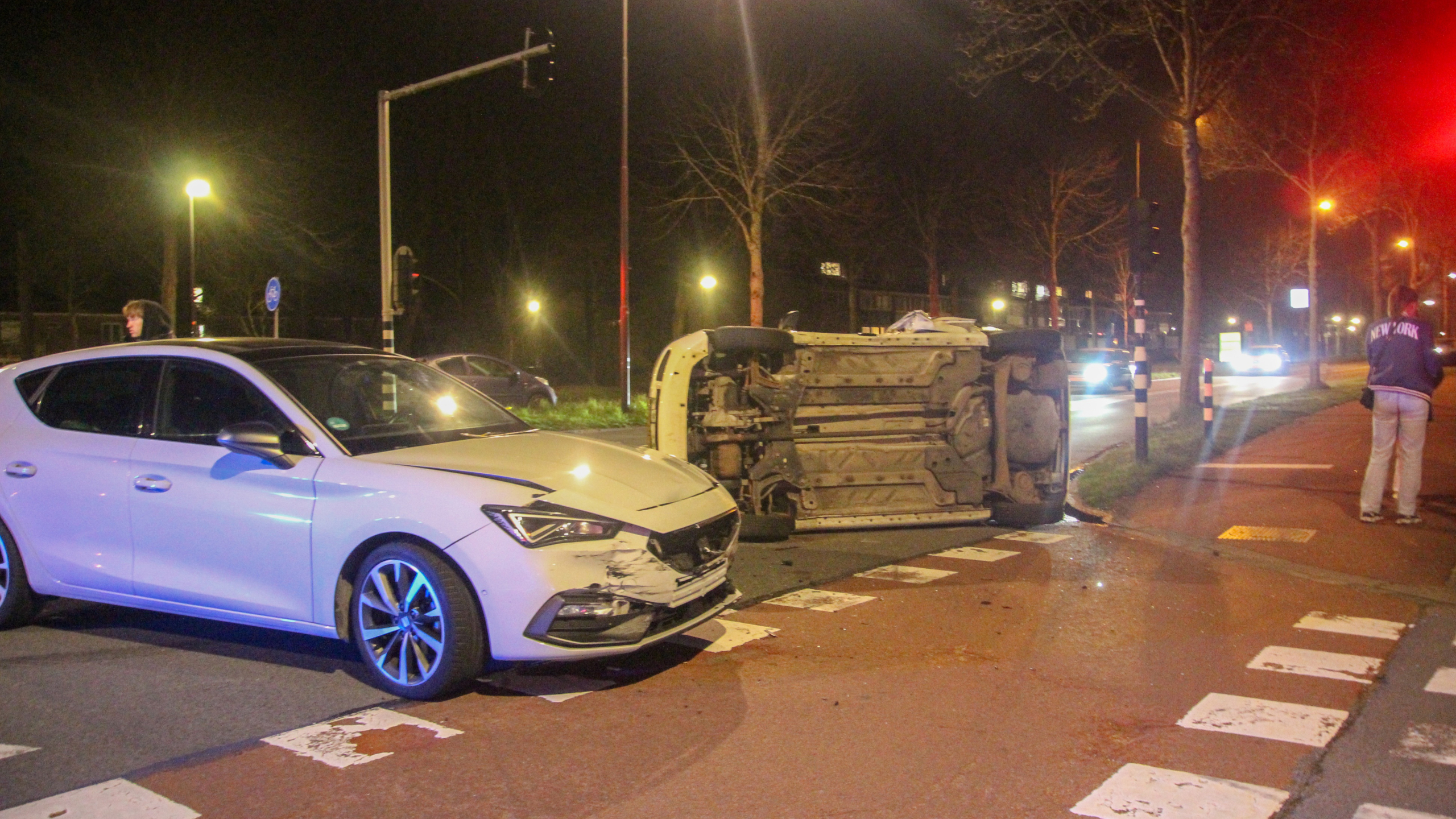 Een witte auto met schade naast een omgevallen voertuig op de weg in een nachtelijke setting, omstanders kijken toe.