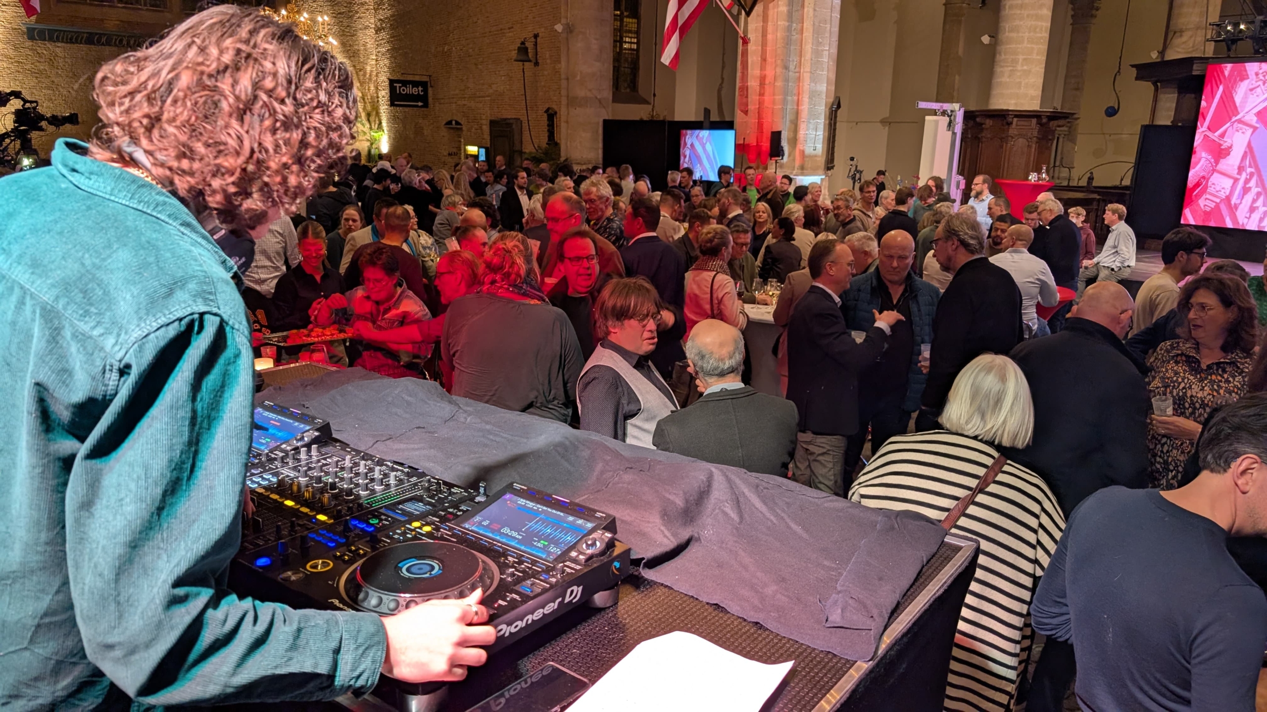 Een DJ draait muziek voor een volle zaal met mensen die staan en met elkaar praten in een grote binnenruimte.