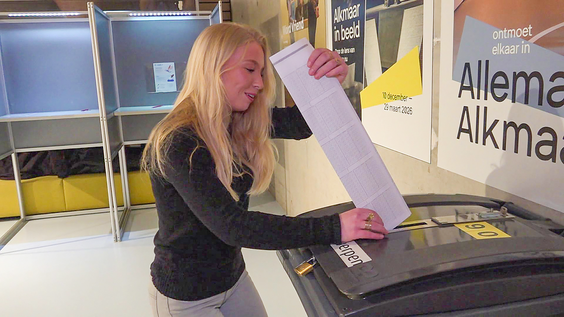 Vrouw deponeert een grote stemlijst in een stembus in een stemlokaal met verkiezingsposters op de achtergrond.