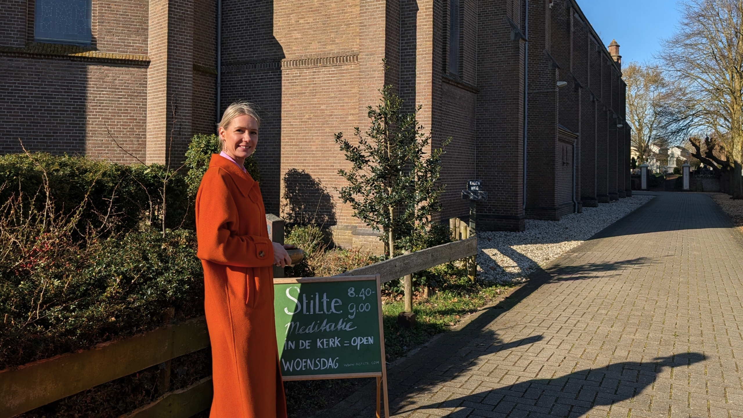 Vrouw in een oranje jas voor een kerk, naast een bord met informatie over meditatie op woensdag.