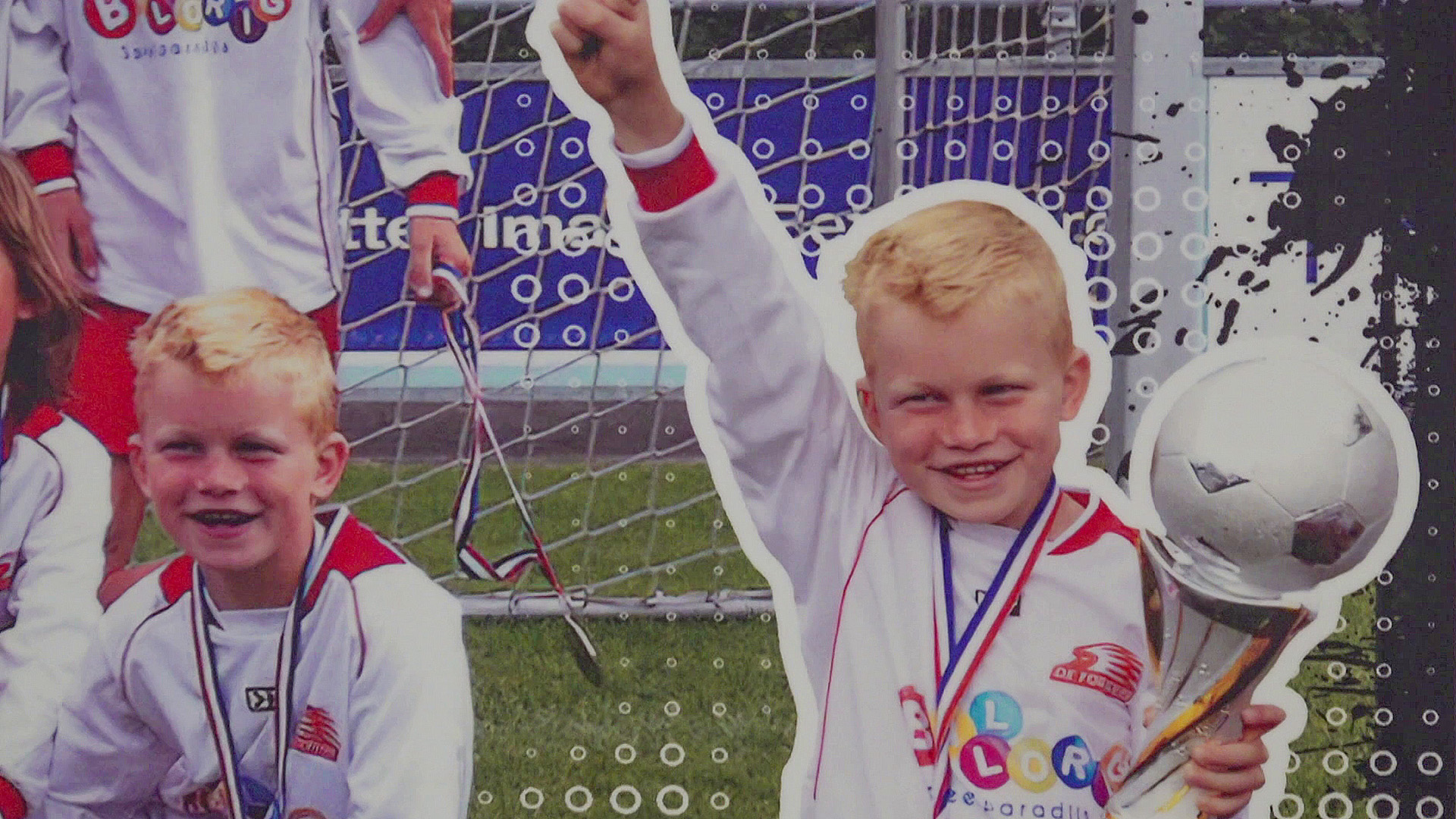 Een kind in een wit sporttenue houdt trots een beker vast naast een voetbaldoel, met een medaille om zijn nek.