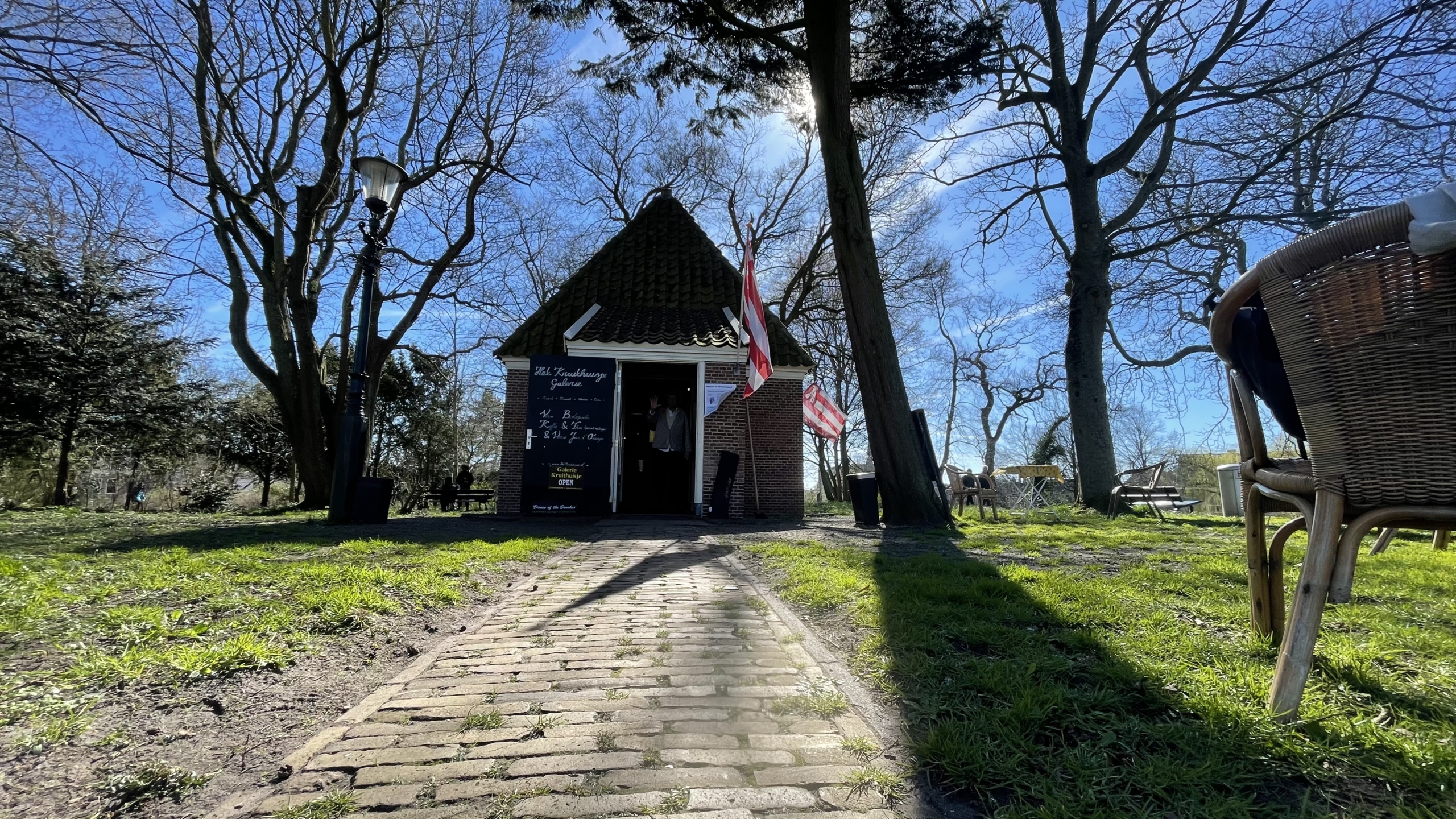 Klein bakstenen gebouwtje met rieten dak in een groen park met kale bomen en een geplaveid pad, naast een tafel en stoel.