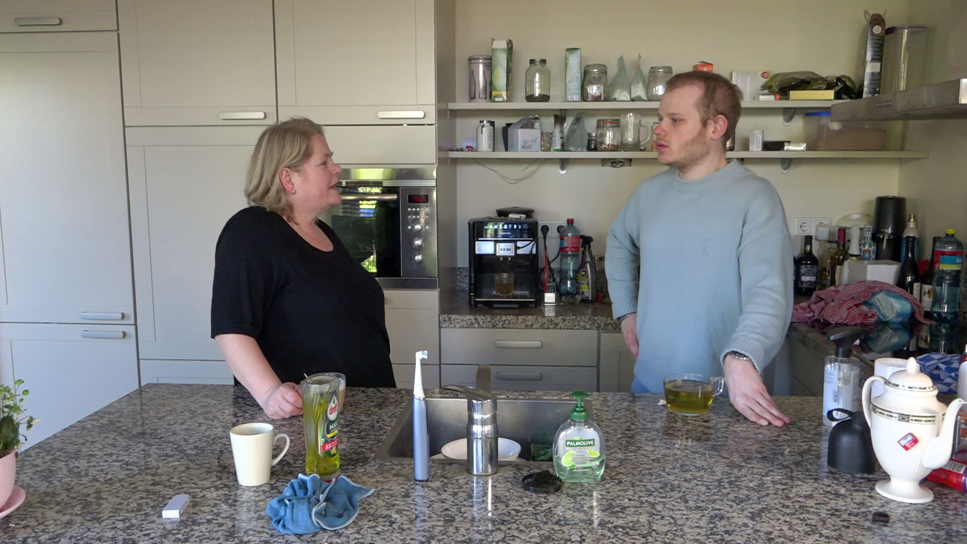 Een man en een vrouw praten met elkaar in een keuken, omringd door keukengerei en voorraadpotten.