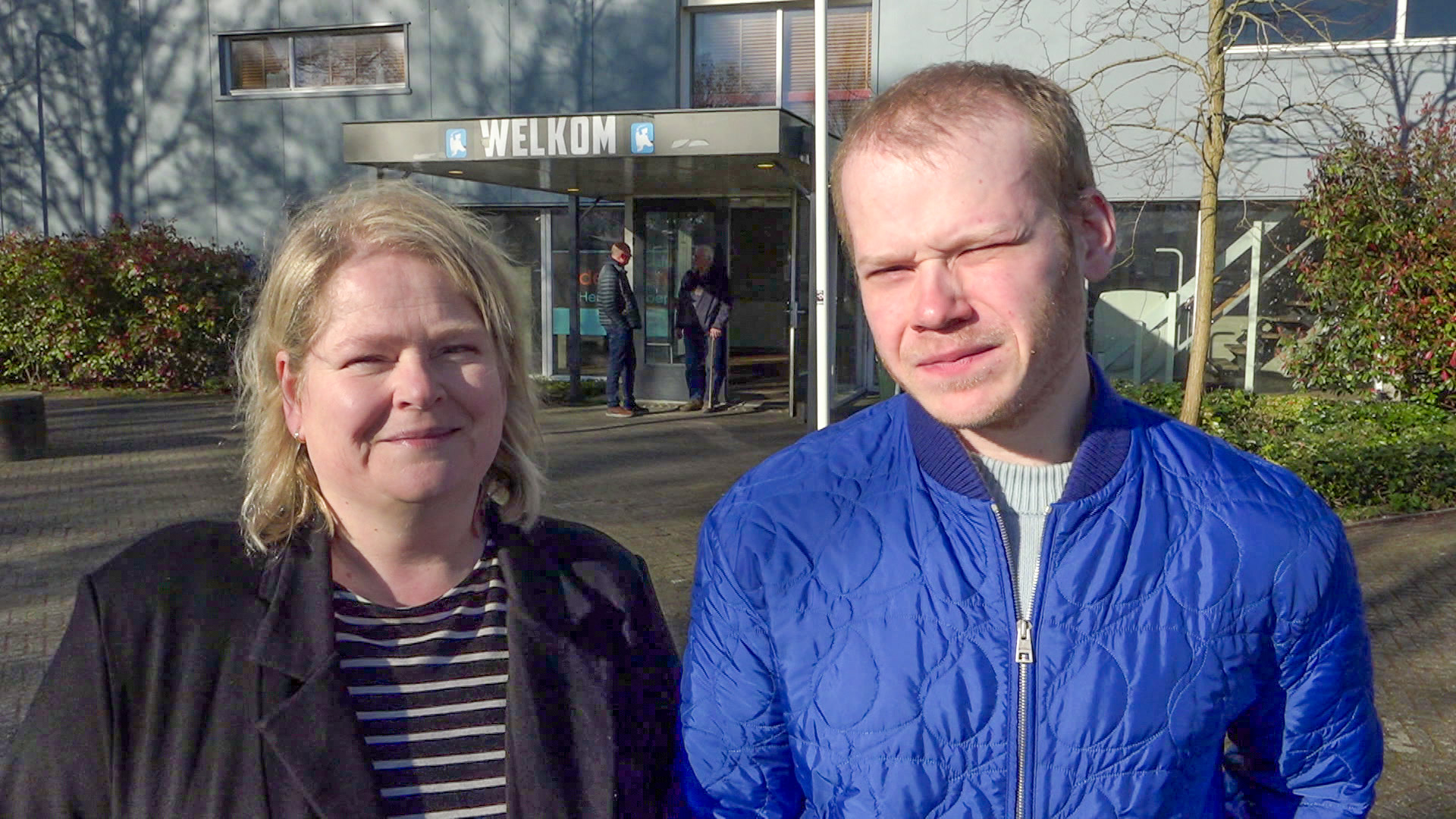 Twee personen staan buiten voor een gebouw met een bord "Welkom".