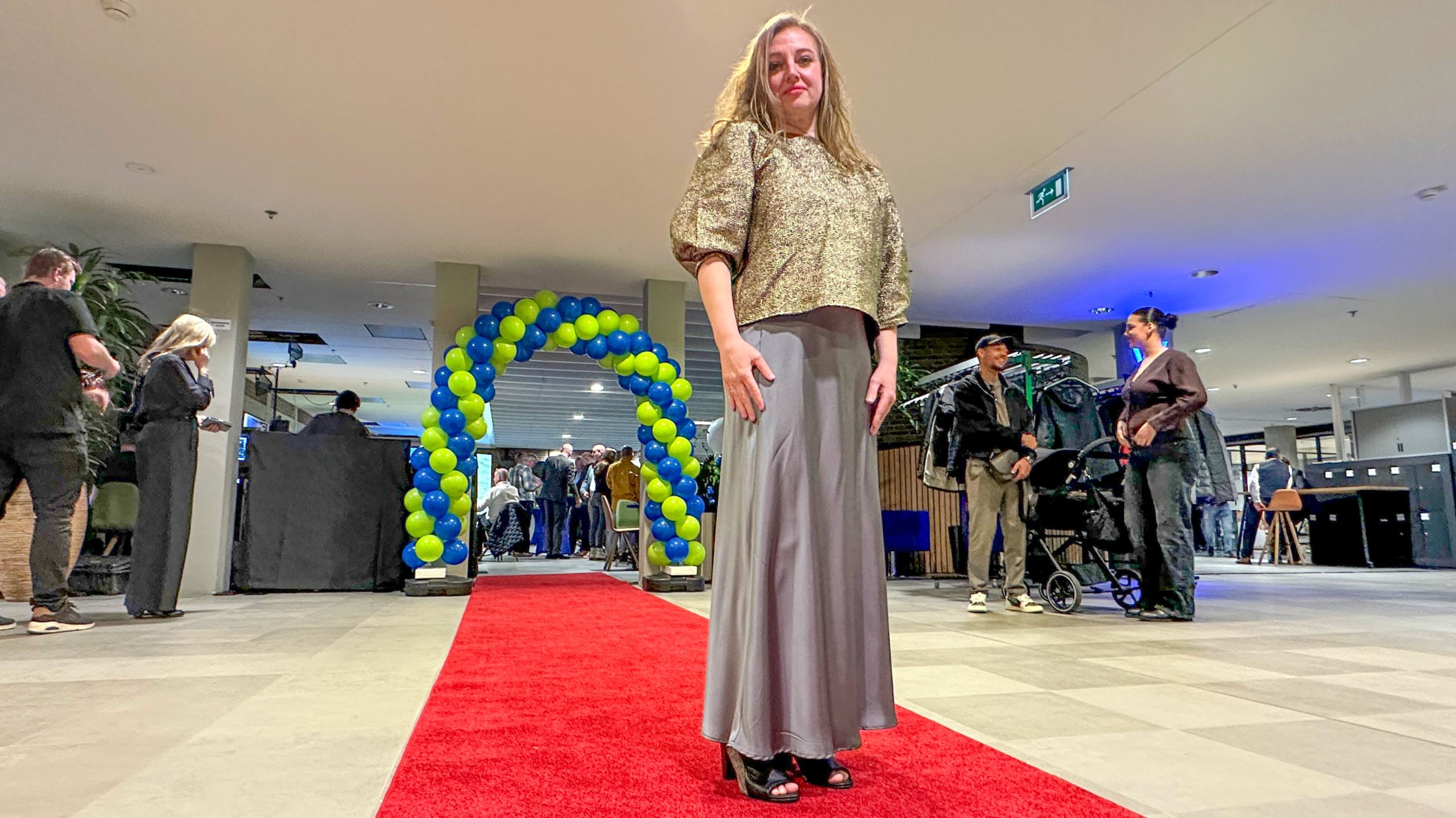 Vrouw in een glinsterende top en lange rok staat op een rode loper, met een boog van blauwe en gele ballonnen op de achtergrond.