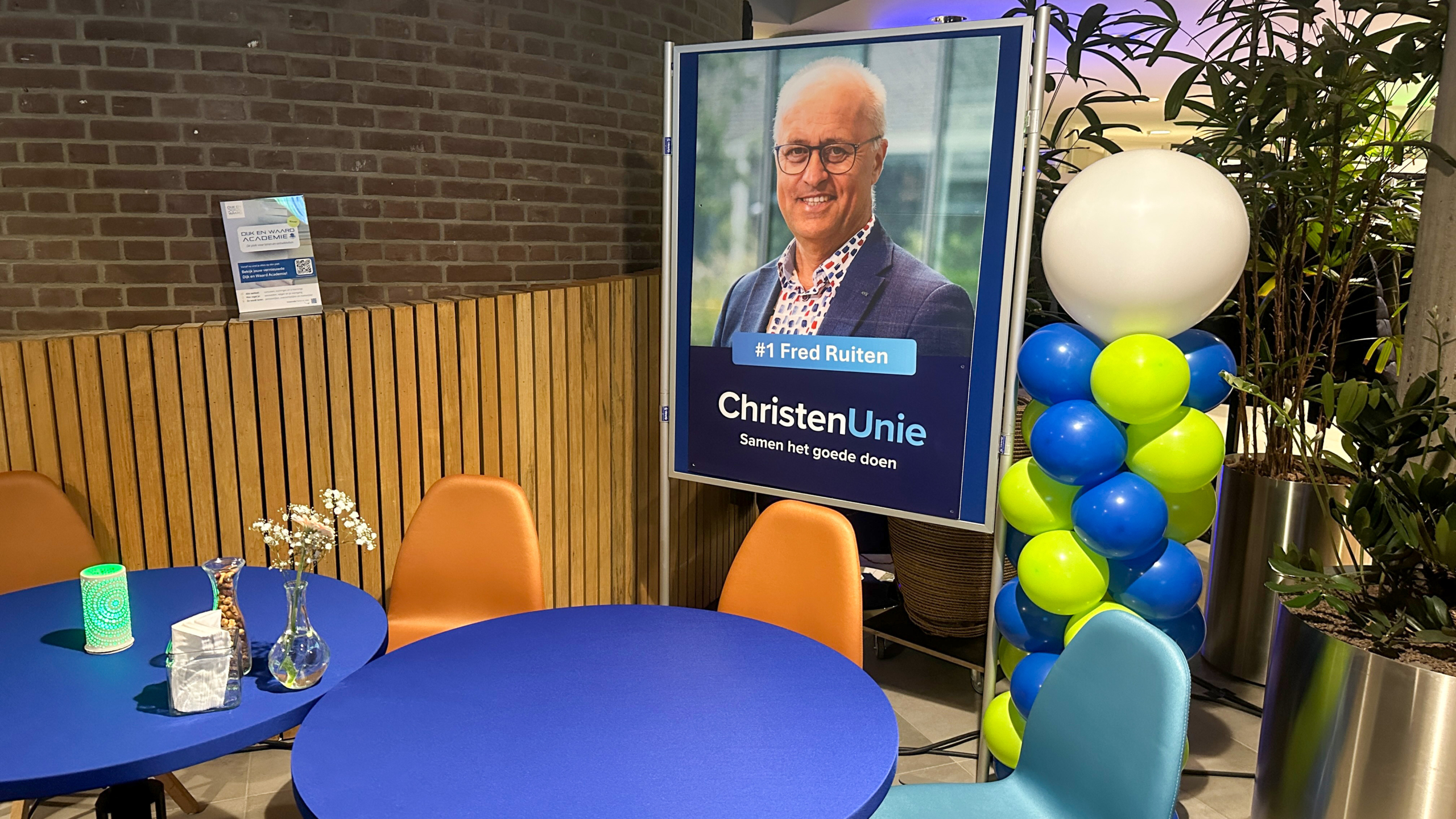 Verkiezingsposter van ChristenUnie met ballondecoratie naast een tafelopstelling in een café-achtige ruimte.