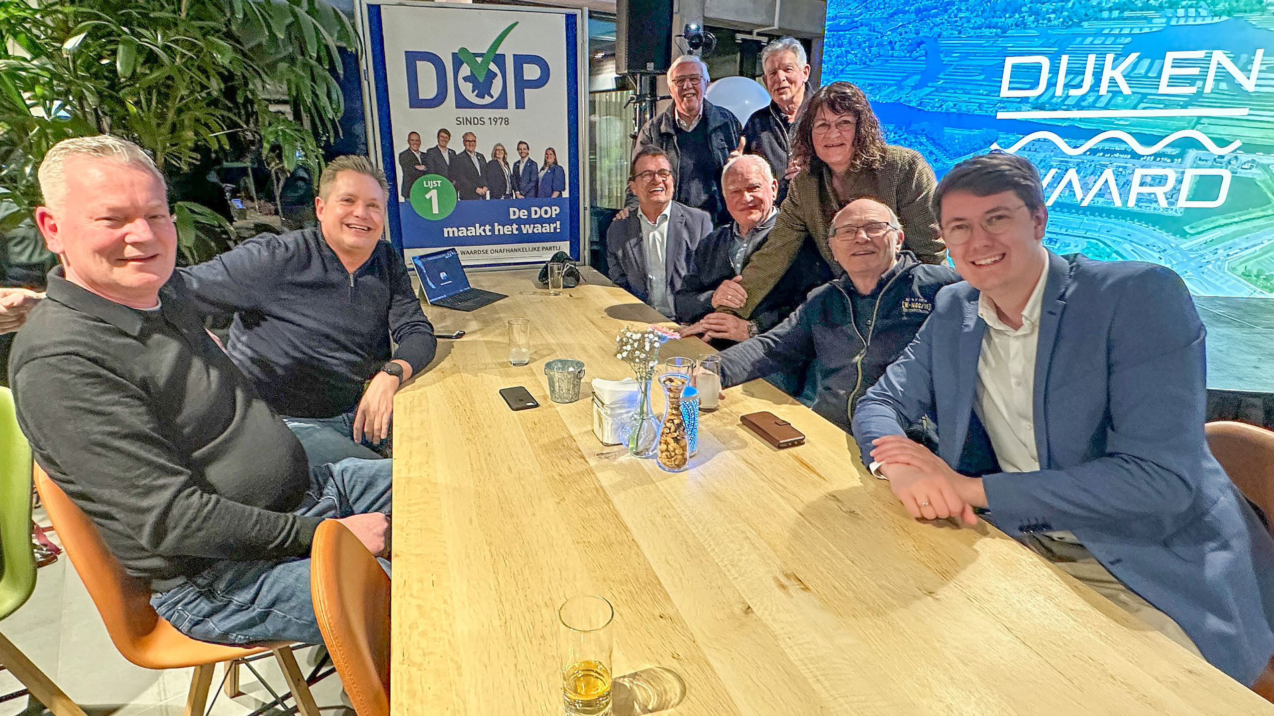 Groep mensen rond een tafel met een poster van de DOP-partij op de achtergrond, in een informele setting.