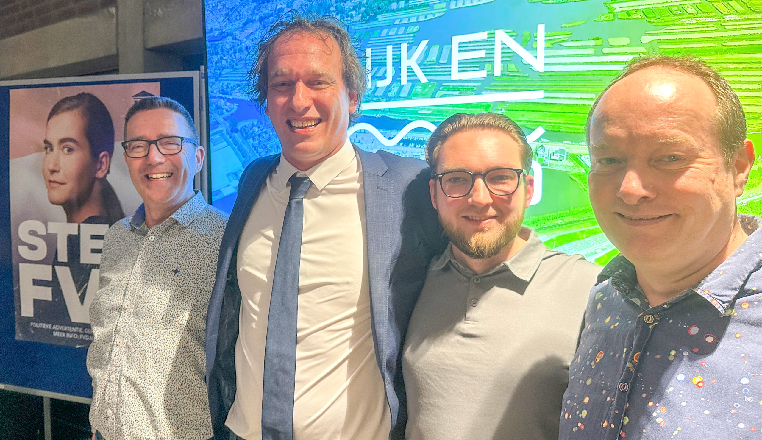 Vier mannen glimlachen naar de camera voor een achtergrond met tekst en een poster van een vrouw met de woorden "STEM FV".