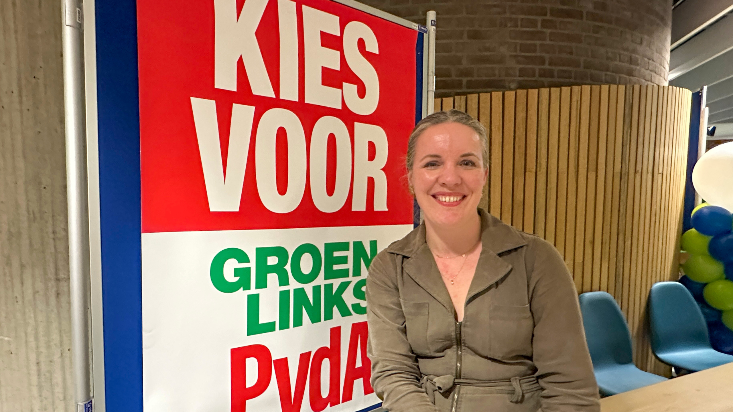 Vrouw glimlacht en zit voor een bord met "Kies voor GroenLinks PvdA" in een binnenruimte met ballonnen.