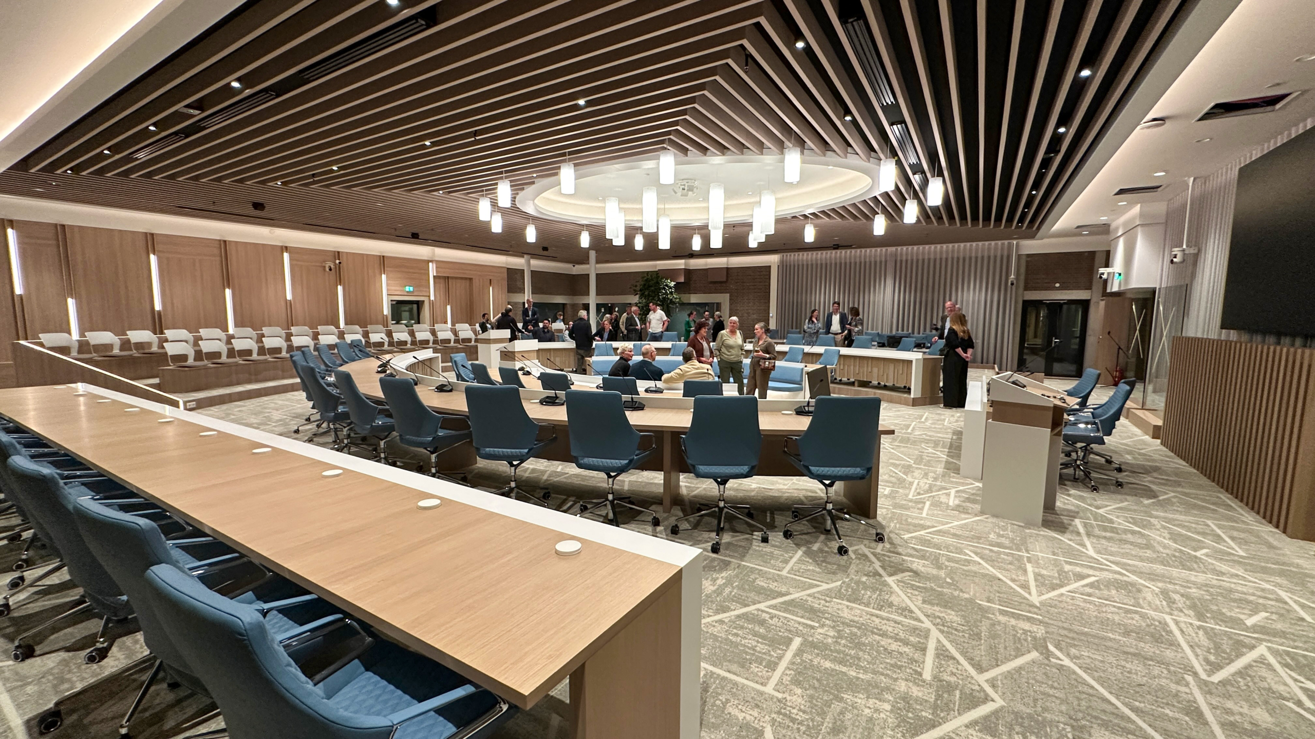 Vergaderruimte met ronde tafels, blauwe stoelen en hanglampen, waar mensen zich informeel verzamelen.