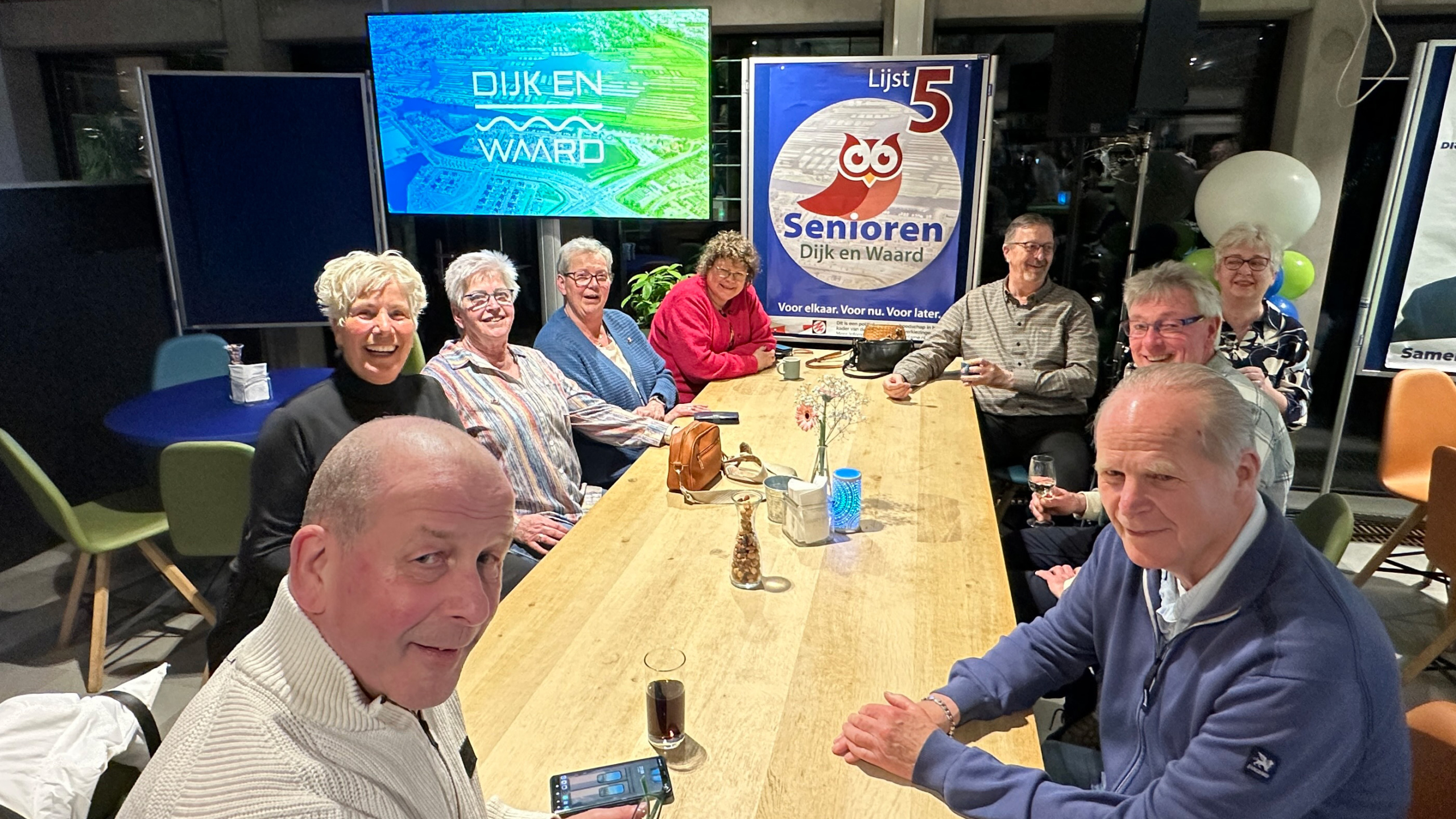 Groep oudere volwassenen aan een tafel in een zaal met een poster voor "Senioren Dijk en Waard Lijst 5" en een scherm met tekst.