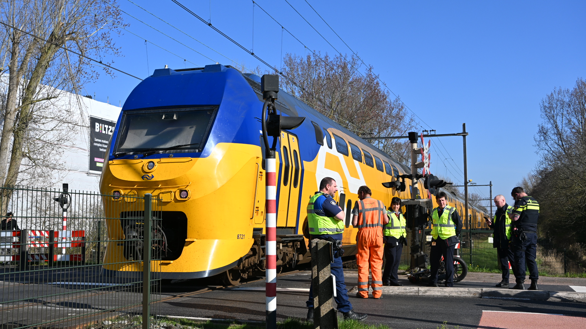 Een gele en blauwe passagierstrein staat bij een spoorwegovergang met politie- en spoormedewerkers op de voorgrond.