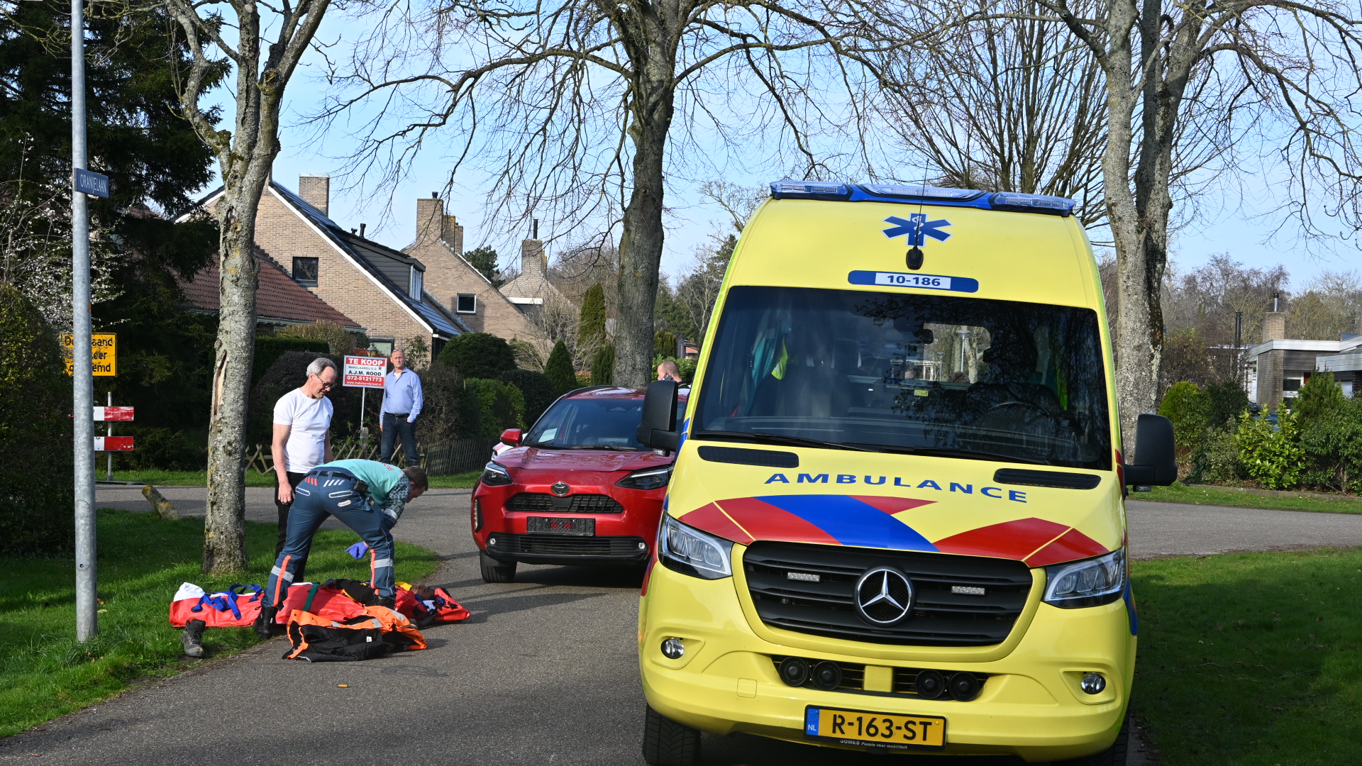Een ambulance staat geparkeerd naast een rode auto op een straat, terwijl twee mensen zich buigen over spullen op de grond.