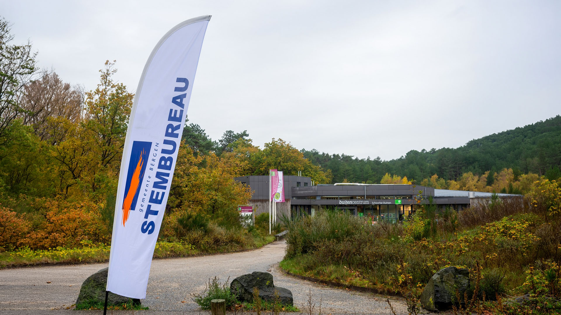 Een stembureauvlag van de gemeente Bergen bij een gebouw omgeven door herfstbomen en een bosrijke omgeving.
