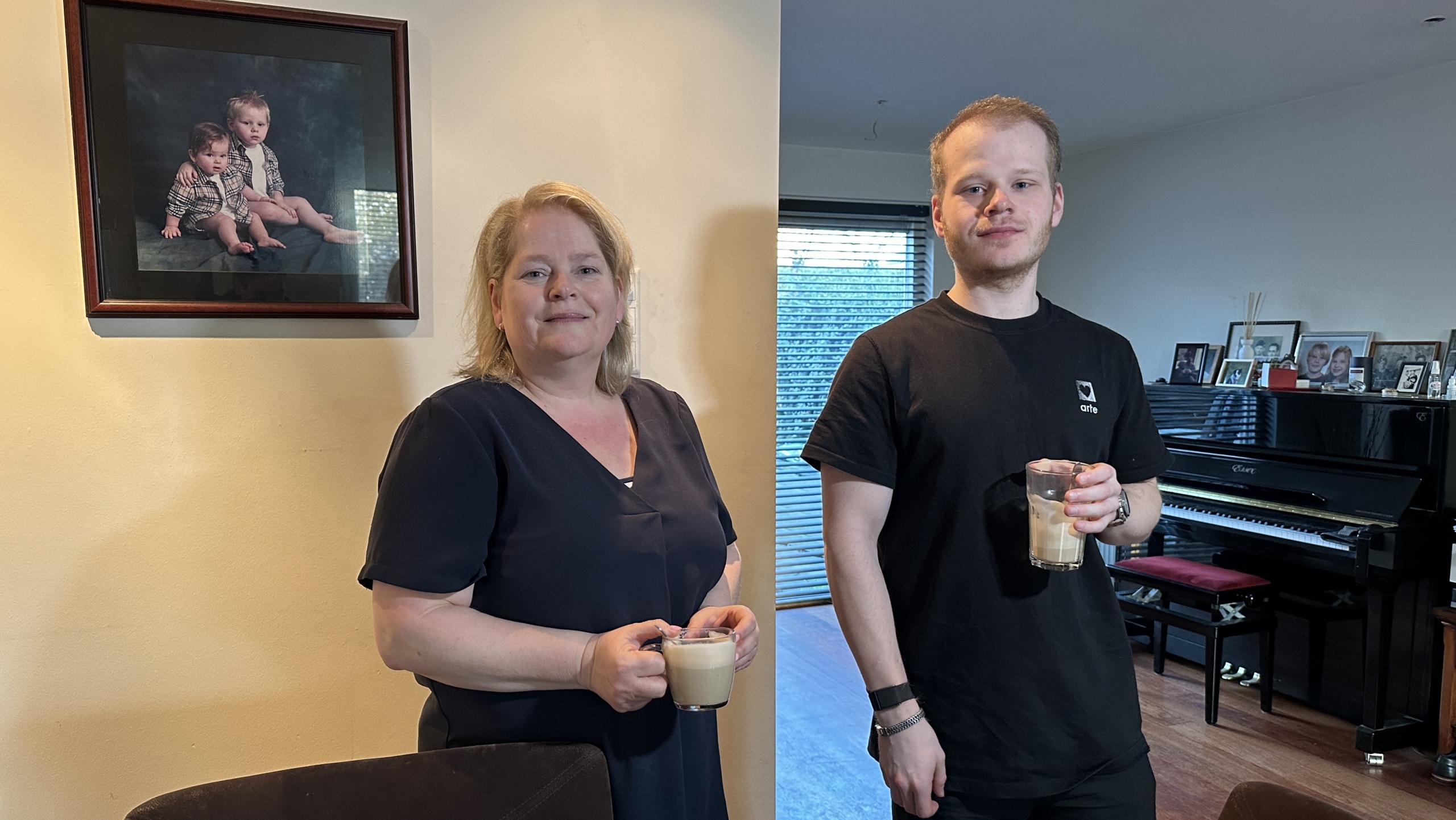 Een vrouw en een man staan in een kamer met een piano, beide houden een glas drinken vast.