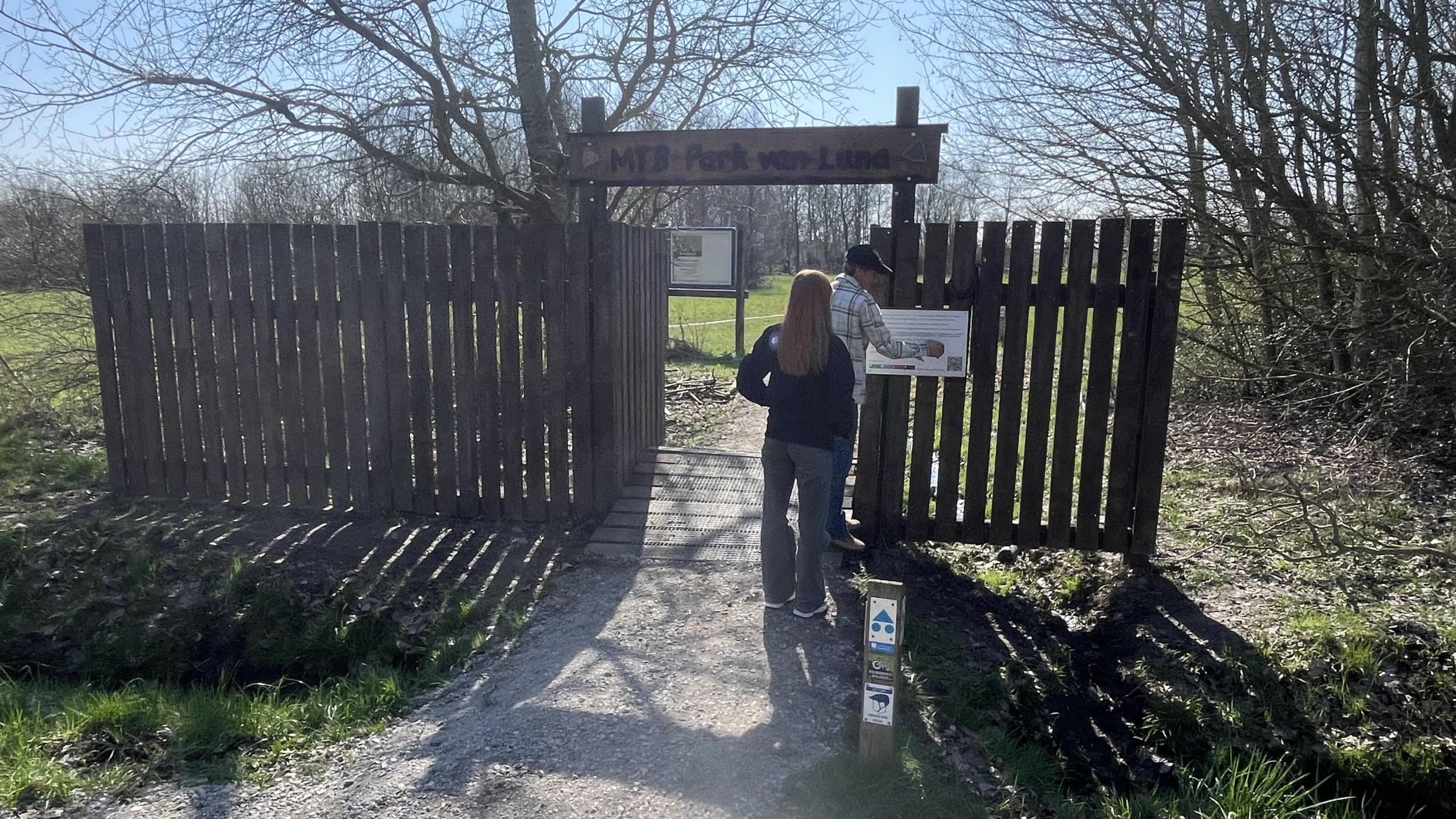 Twee mensen staan voor de ingang van MTB-Park van Luna, omgeven door bomen en een grasveld.