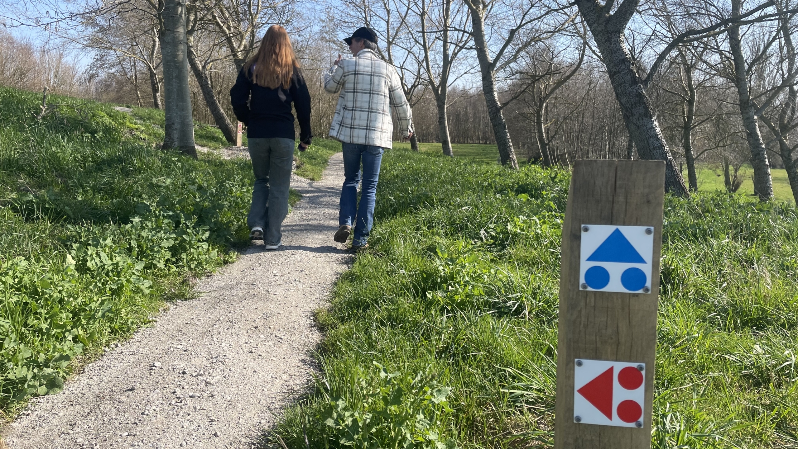 Twee mensen wandelen over een pad door een park met bomen; een paaltje met routeborden is zichtbaar.
