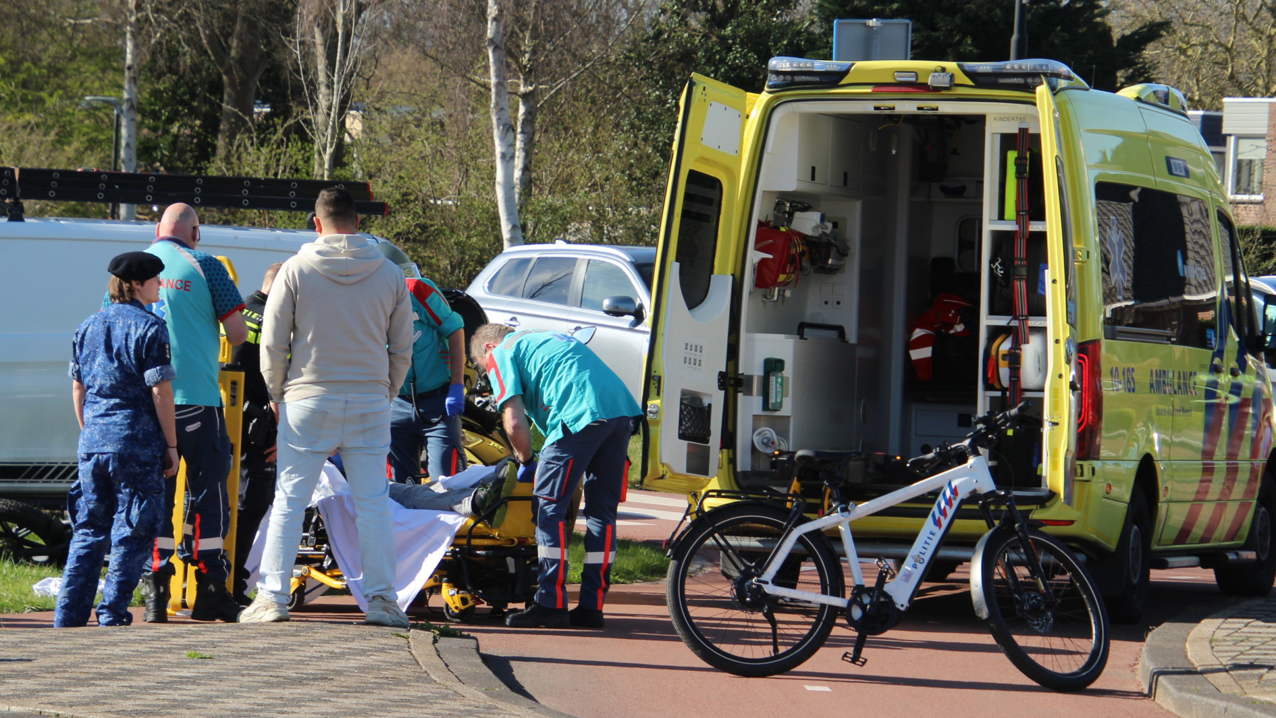 Mensen helpen een persoon op een brancard bij een ambulance, met een geopende achterdeur in een stedelijke omgeving.